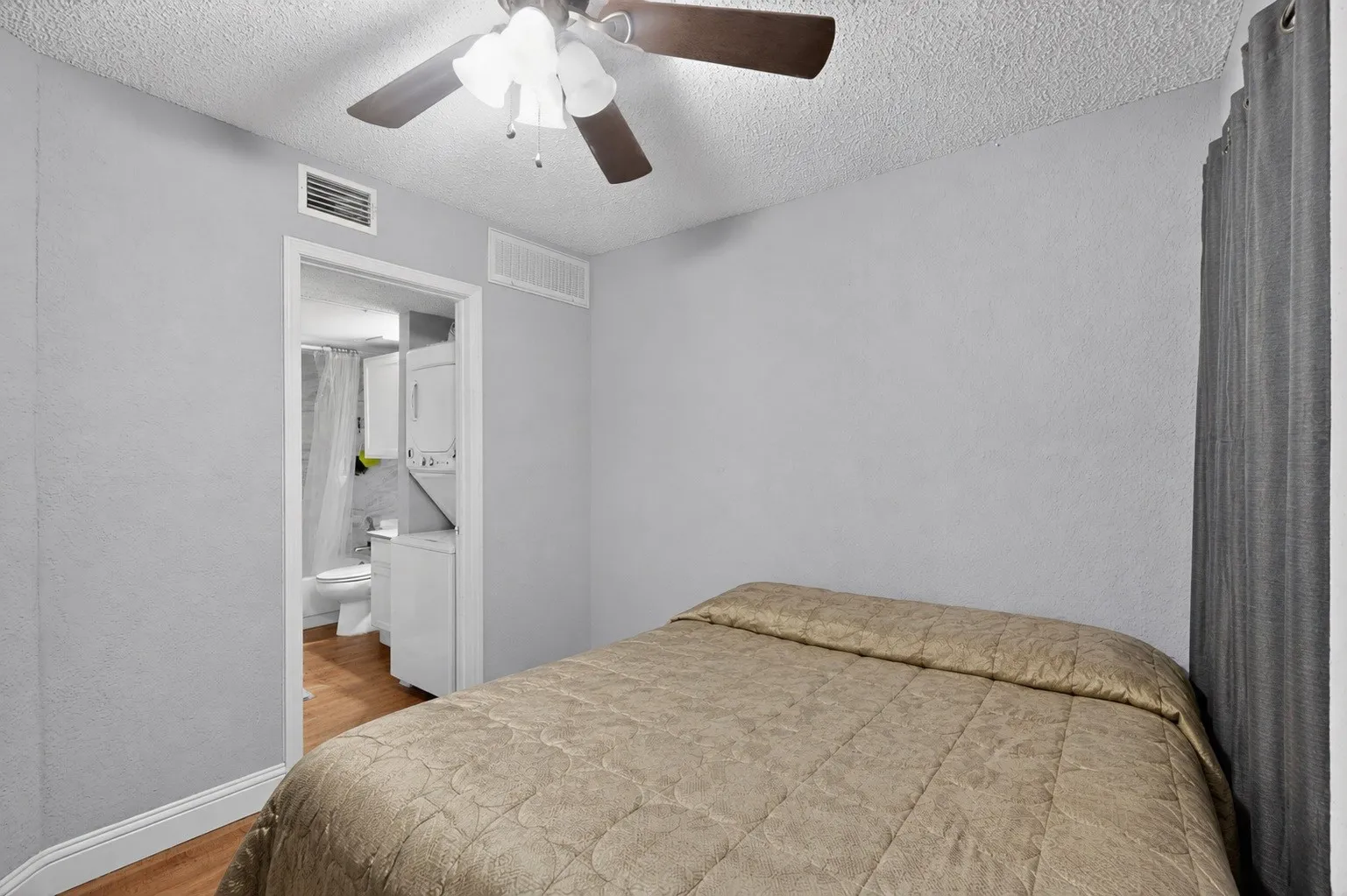 Bedroom featuring a ceiling fan with light kit, textured ceiling, and a doorway opening to a bathroom with wood-finish flooring and a stacked washer and dryer unit