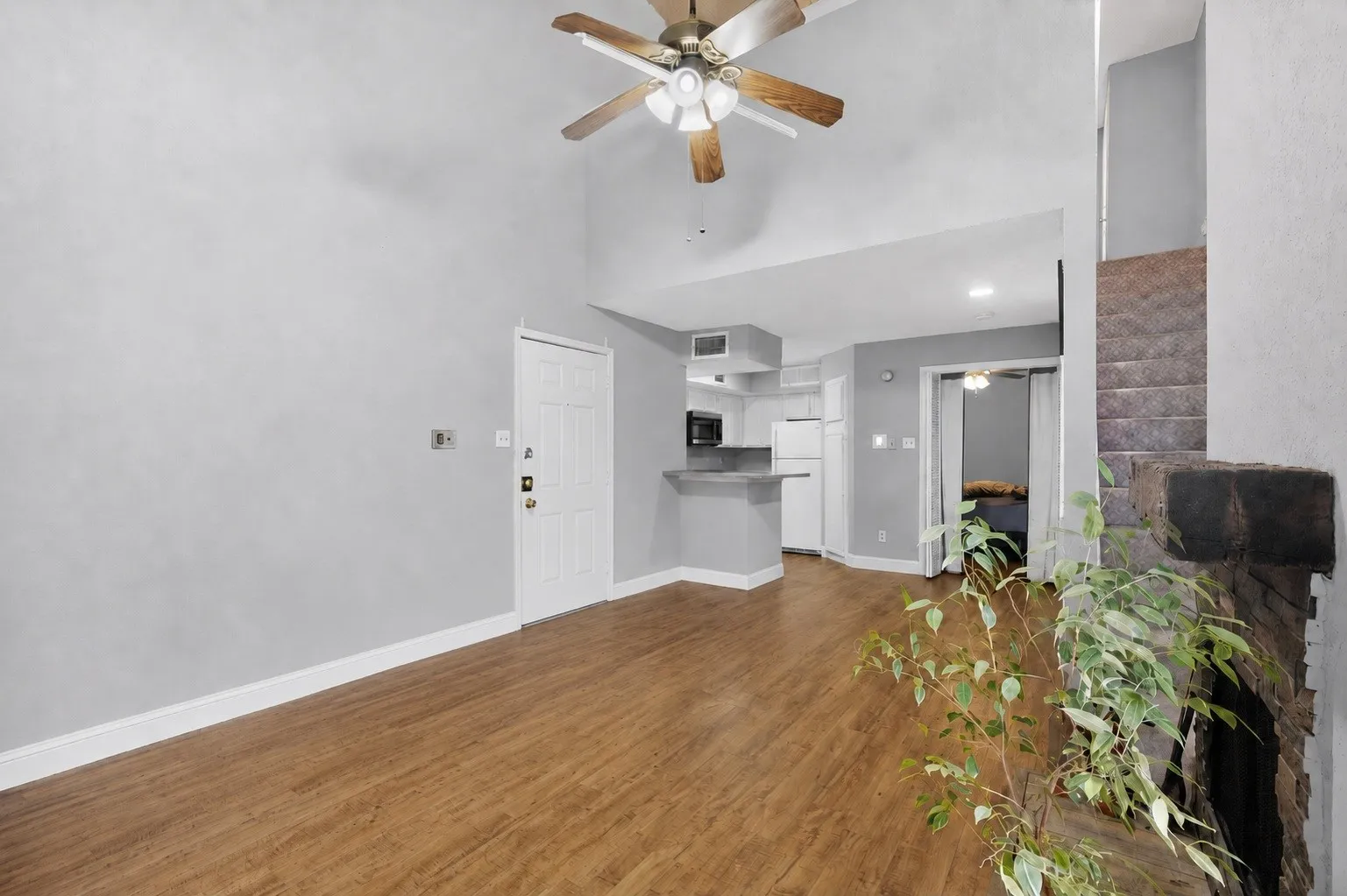 Vaulted ceiling with a ceiling fan, wood-finish flooring, a kitchen area with white cabinetry, and a stone fireplace surround