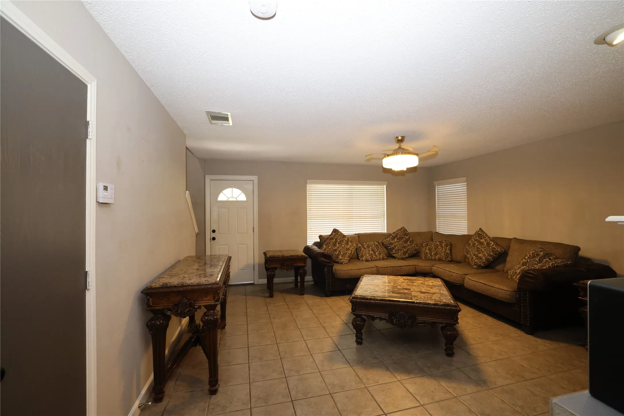 Spacious living area featuring tile flooring, neutral wall tones, multiple windows with blinds, a ceiling fan with integrated lighting, and a white entry door with an arched glass insert