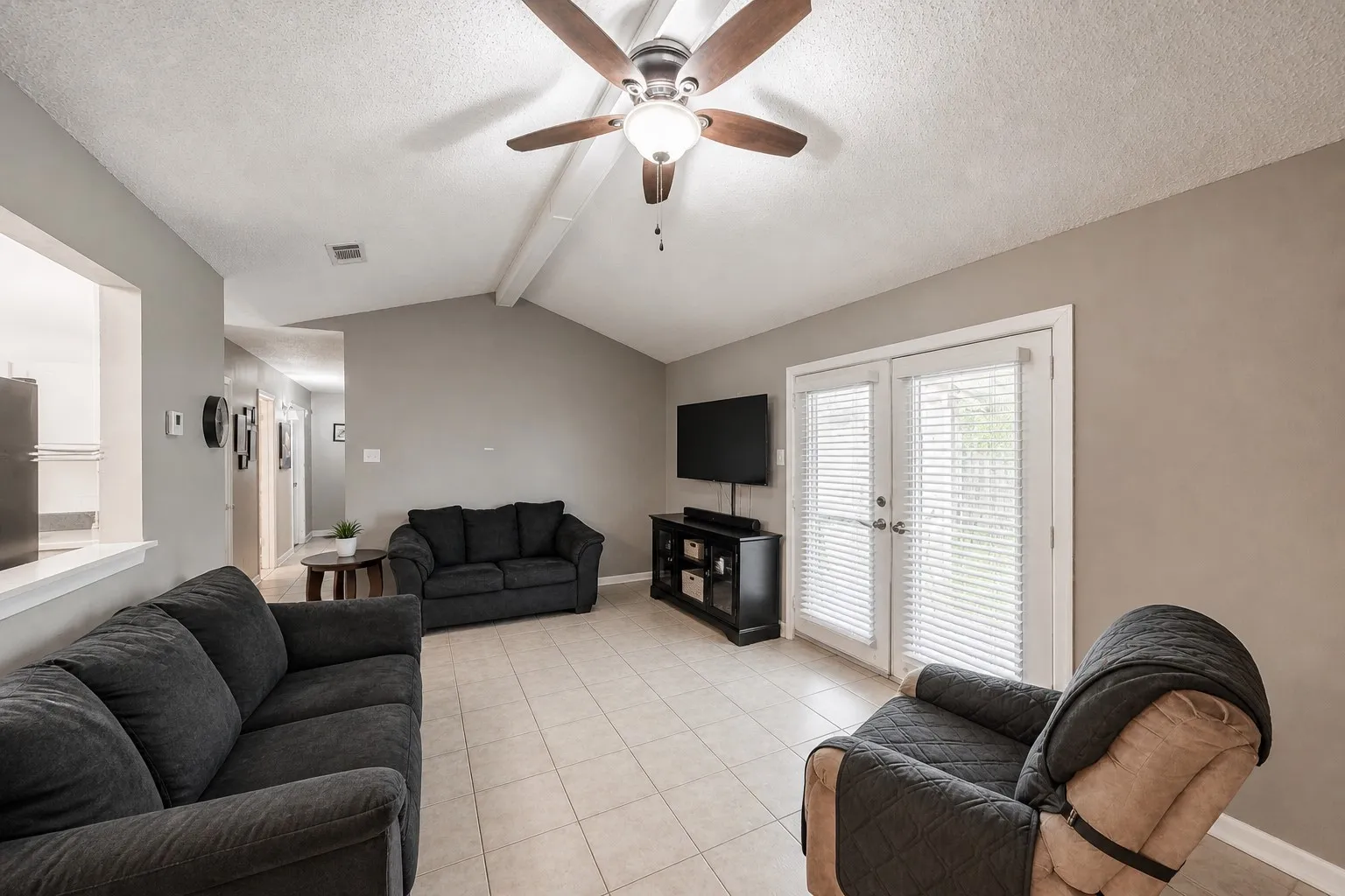 Spacious living area featuring vaulted ceilings, tile flooring, and a ceiling fan. French doors lead to covered patio & backyard.  Photo has been digitally enhanced.