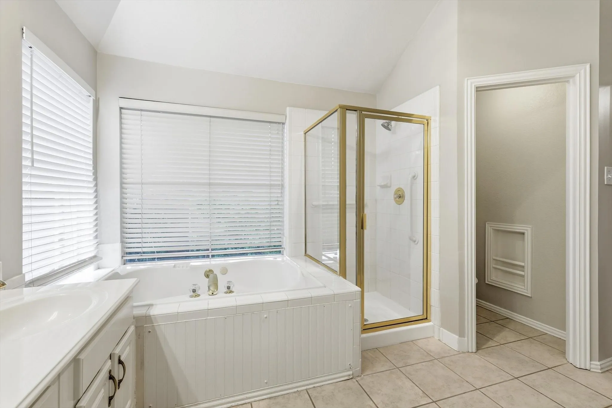 Neutral-toned bathroom featuring a built-in tub with tile surround, a separate shower with a gold-framed glass enclosure, and a vanity with a white countertop