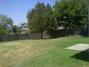 Spacious backyard featuring a large grass area, mature trees, a wood fence perimeter, and a concrete patio slab