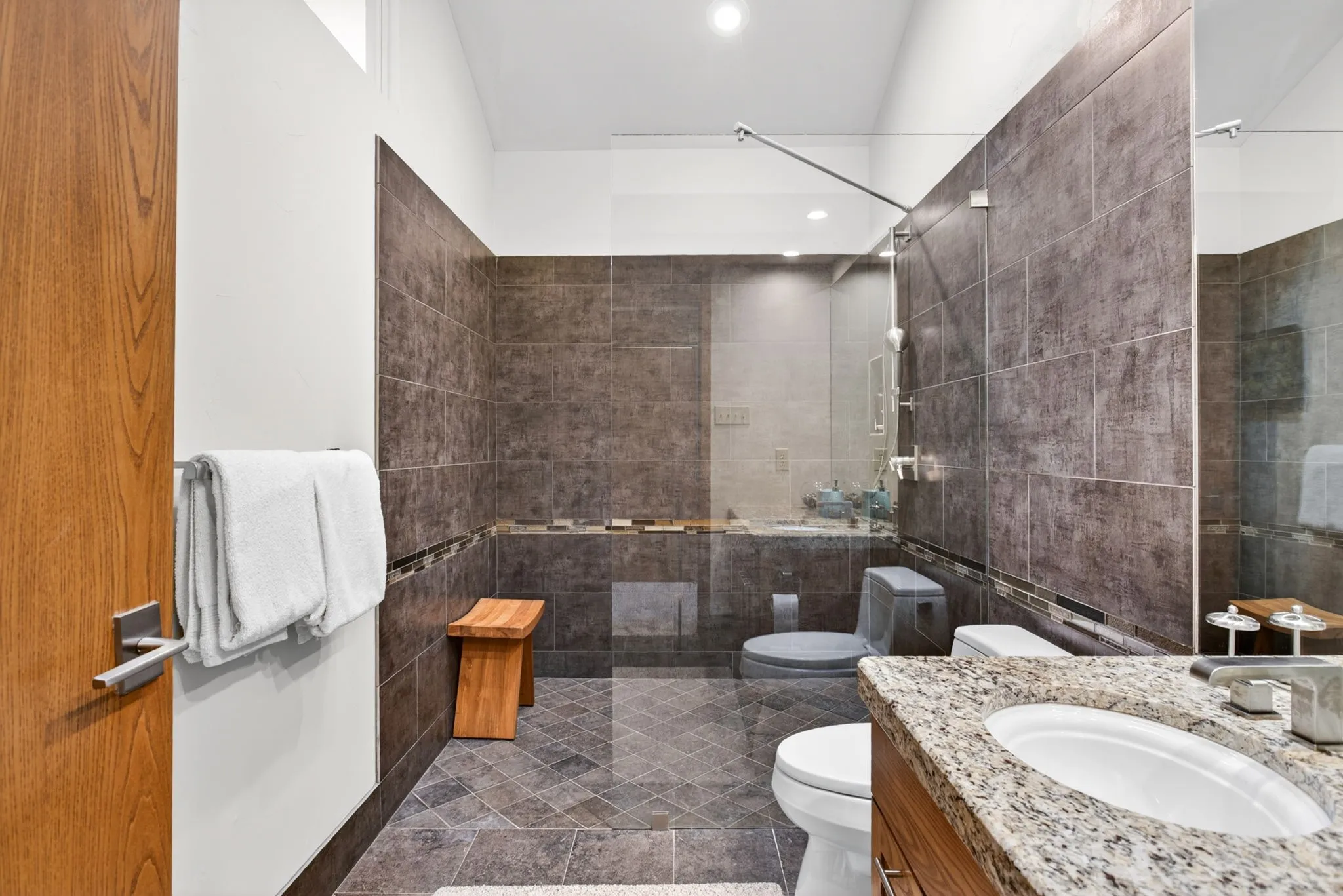 Bathroom featuring a granite countertop vanity with an undermount sink, a glass-enclosed shower with dark tile surround, and coordinating dark tile flooring