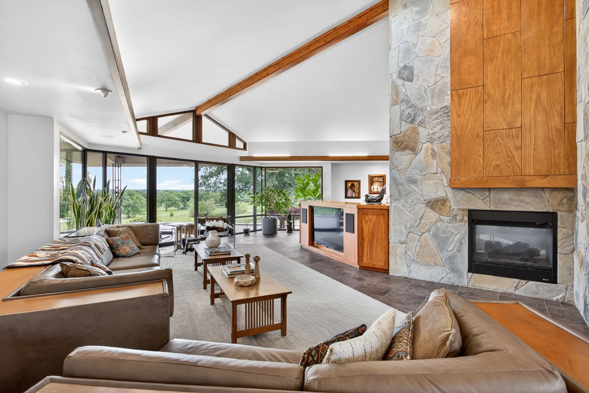 Expansive living area featuring a stone fireplace with a wood surround, vaulted ceilings with exposed wood beams, and a wall of windows offering landscape views
