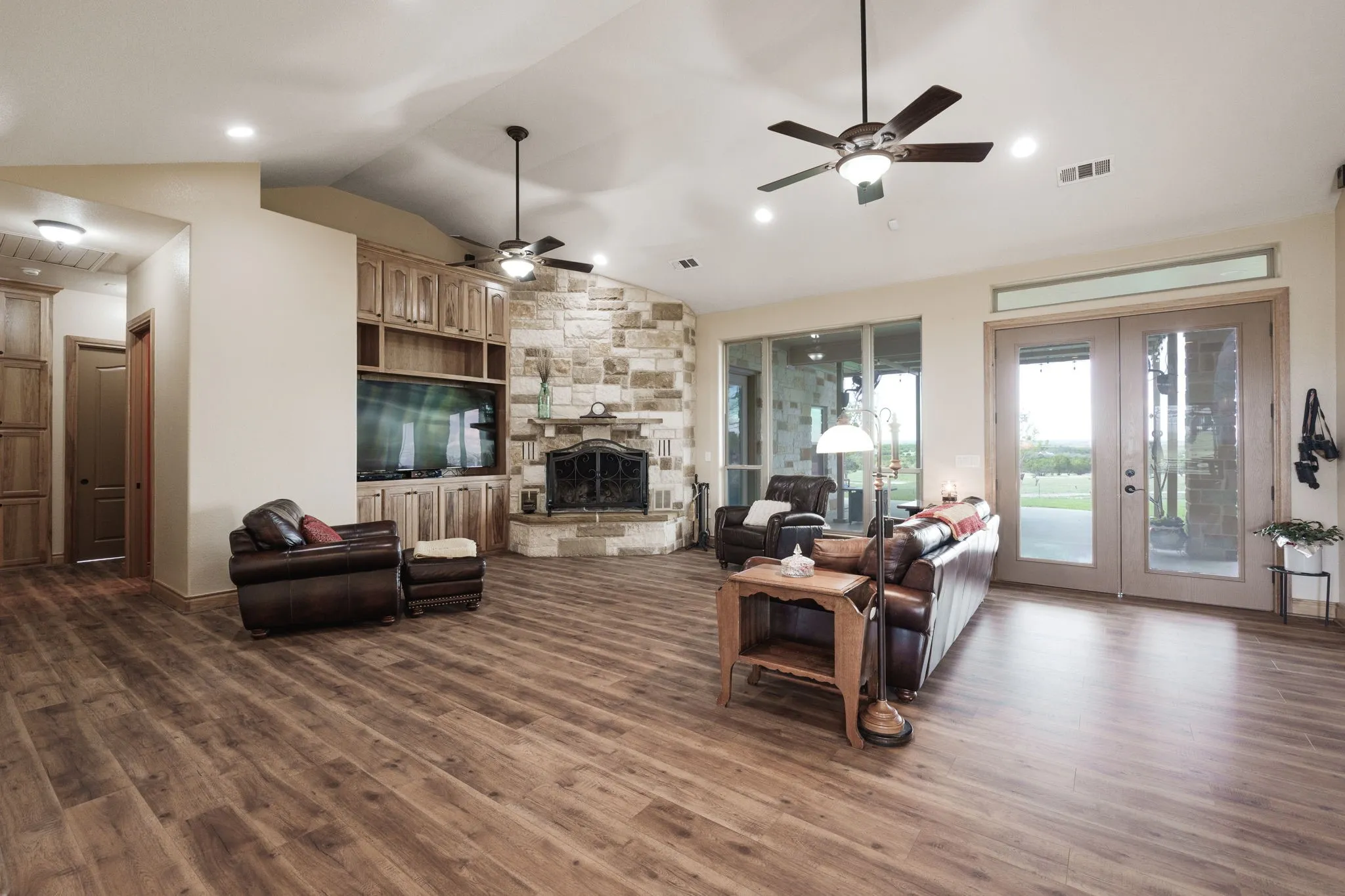 Expansive living space featuring wood-finish flooring, a stone-clad fireplace, integrated wood cabinetry, a vaulted ceiling with recessed lighting, and glass panel doors leading to the exterior