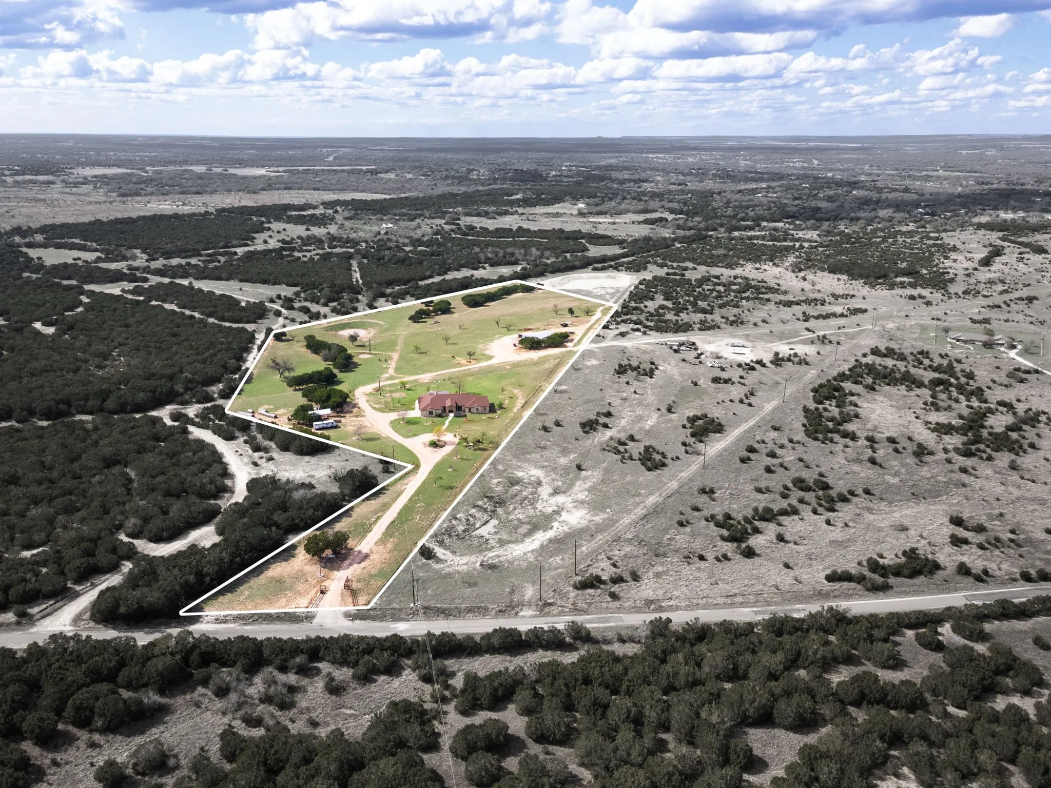 Expansive rural property featuring a main residence with a red tile roof, an unpaved driveway, mature trees, open grassy areas, and surrounding natural brushland