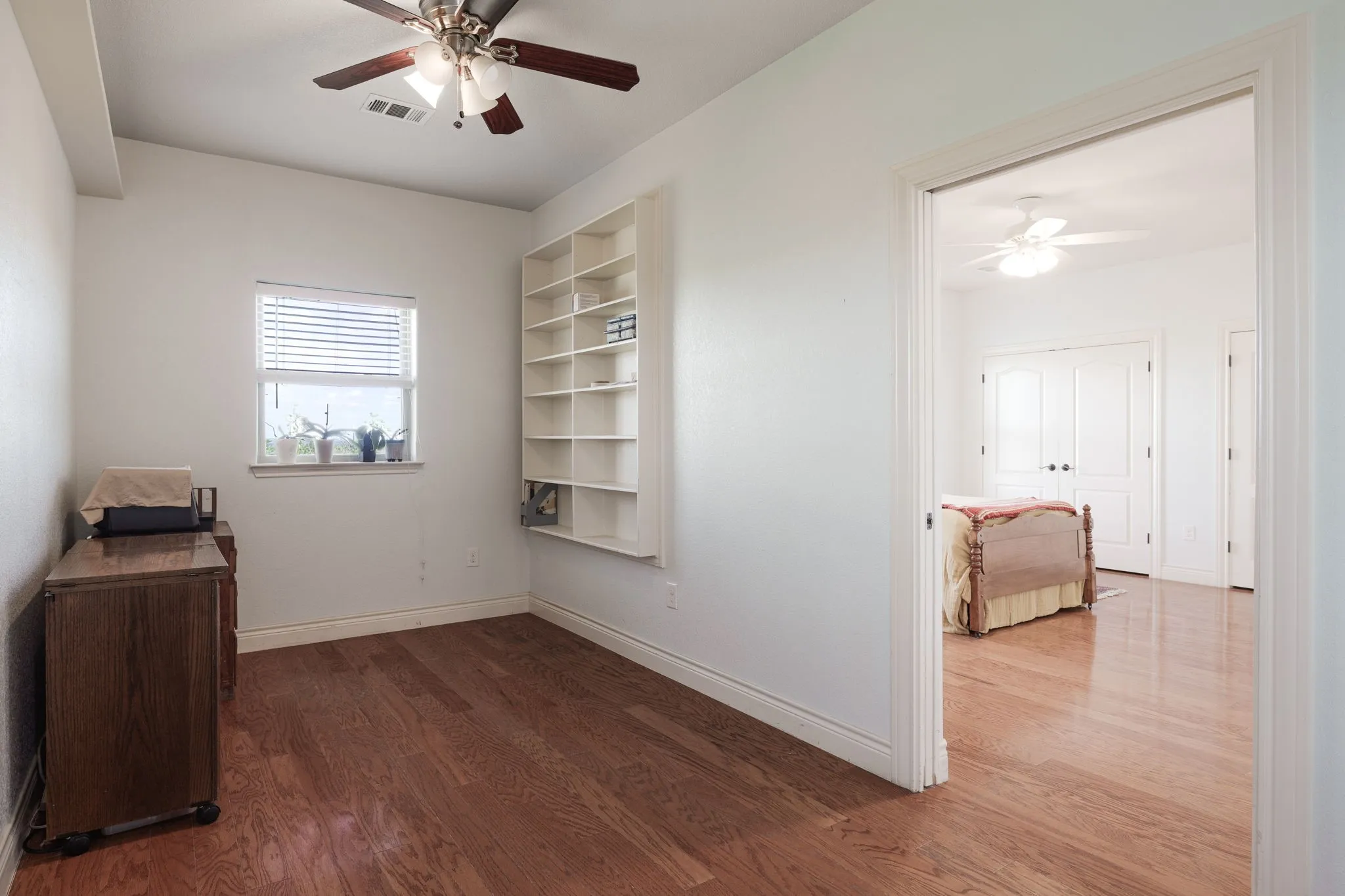 Hardwood-finish flooring throughout, featuring a built-in shelving unit, overhead ceiling fan with lighting, a window with blinds, and white baseboards