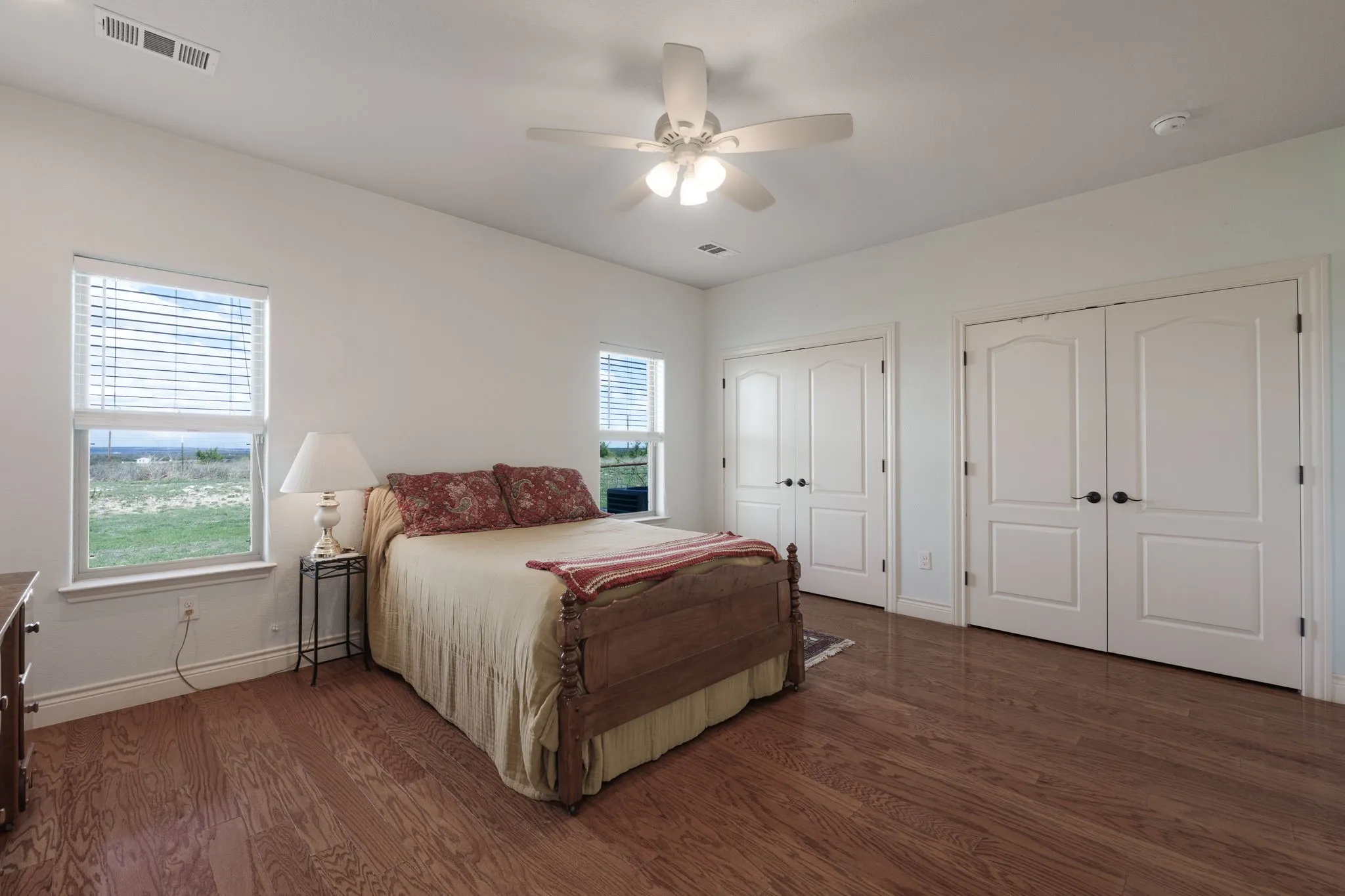 Spacious room featuring wood-finish flooring, two sets of white double-door closets with dark hardware, a ceiling fan with integrated lighting, and two windows equipped with blinds