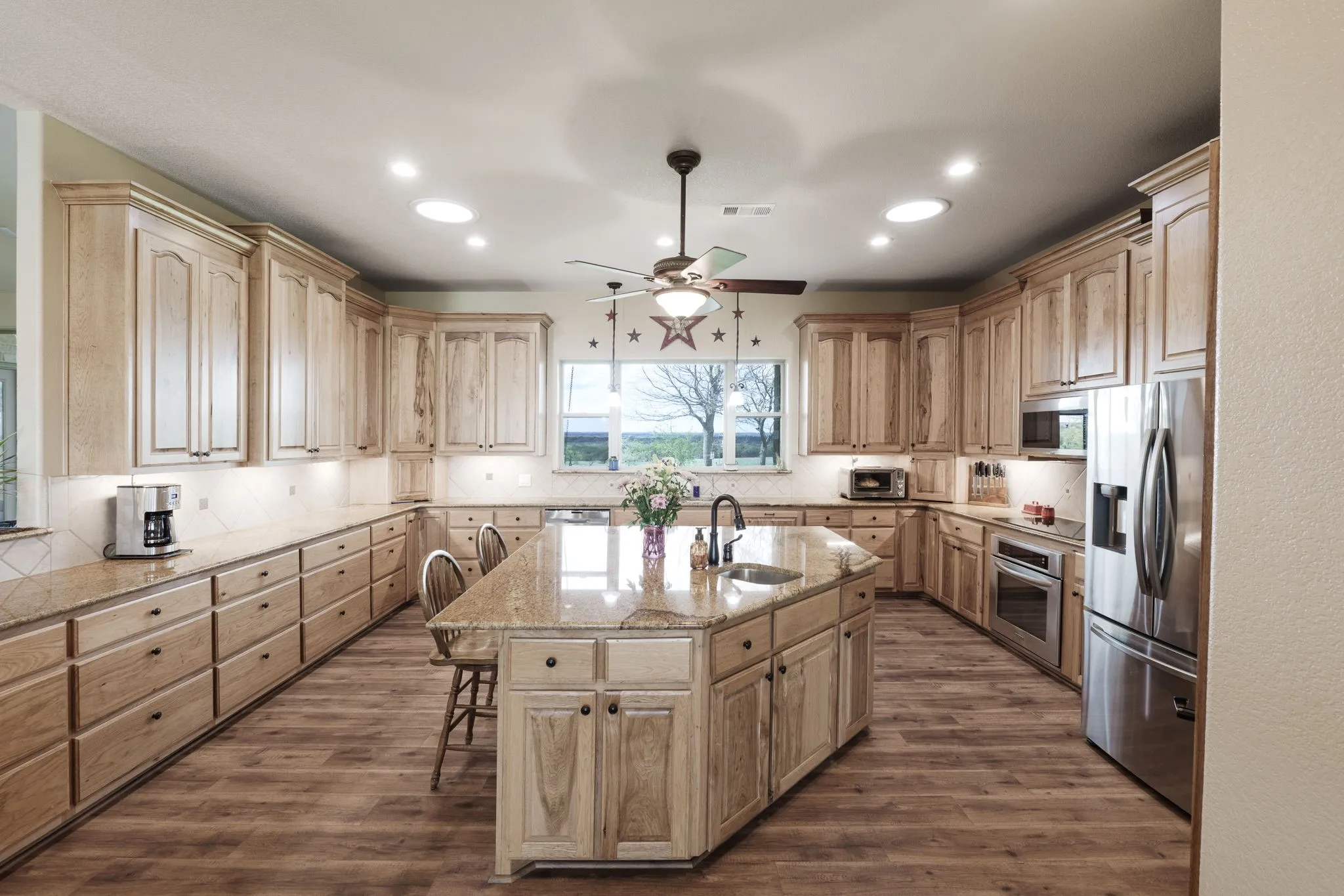 Expansive kitchen featuring wood-finish flooring, abundant light-toned cabinetry, a large center island with a built-in sink, stone countertops, and stainless steel appliances