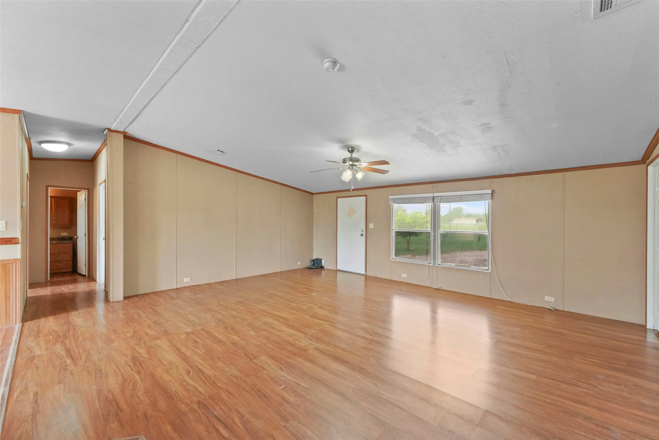 Spacious living area featuring wood-finish flooring, neutral wall tones, a ceiling fan, and prominent window with outdoor scenery