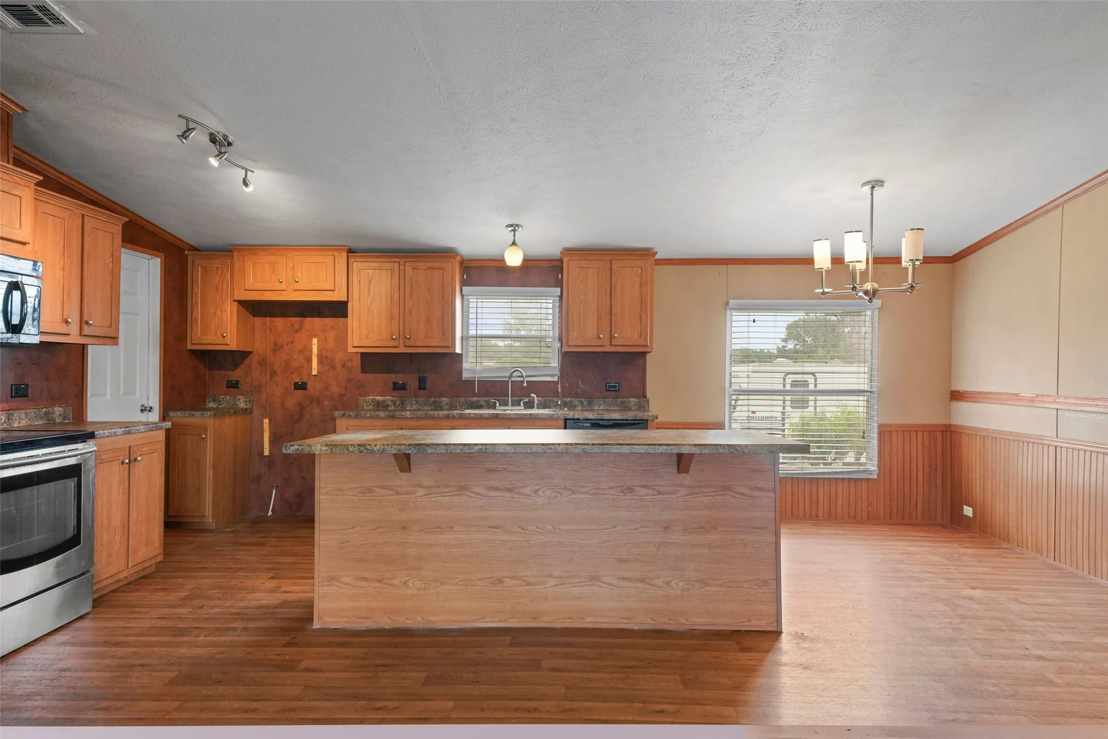 Open-concept kitchen and dining area featuring wood-finish flooring, wood cabinetry, stainless steel appliances, and a central island with a countertop extension