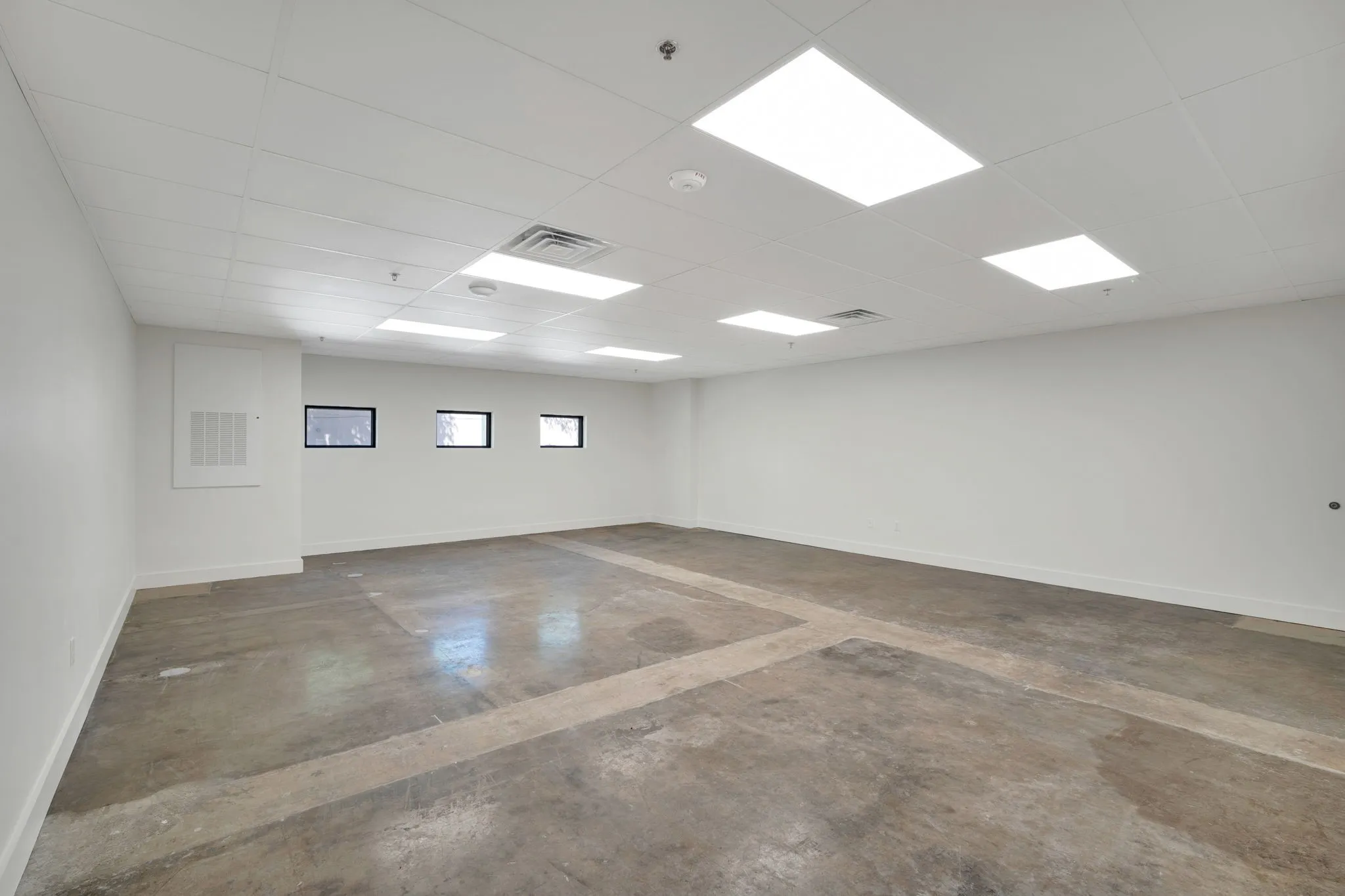 Spacious room featuring concrete flooring, a grid ceiling with integrated rectangular panel lighting, and three rectangular windows with dark frames
