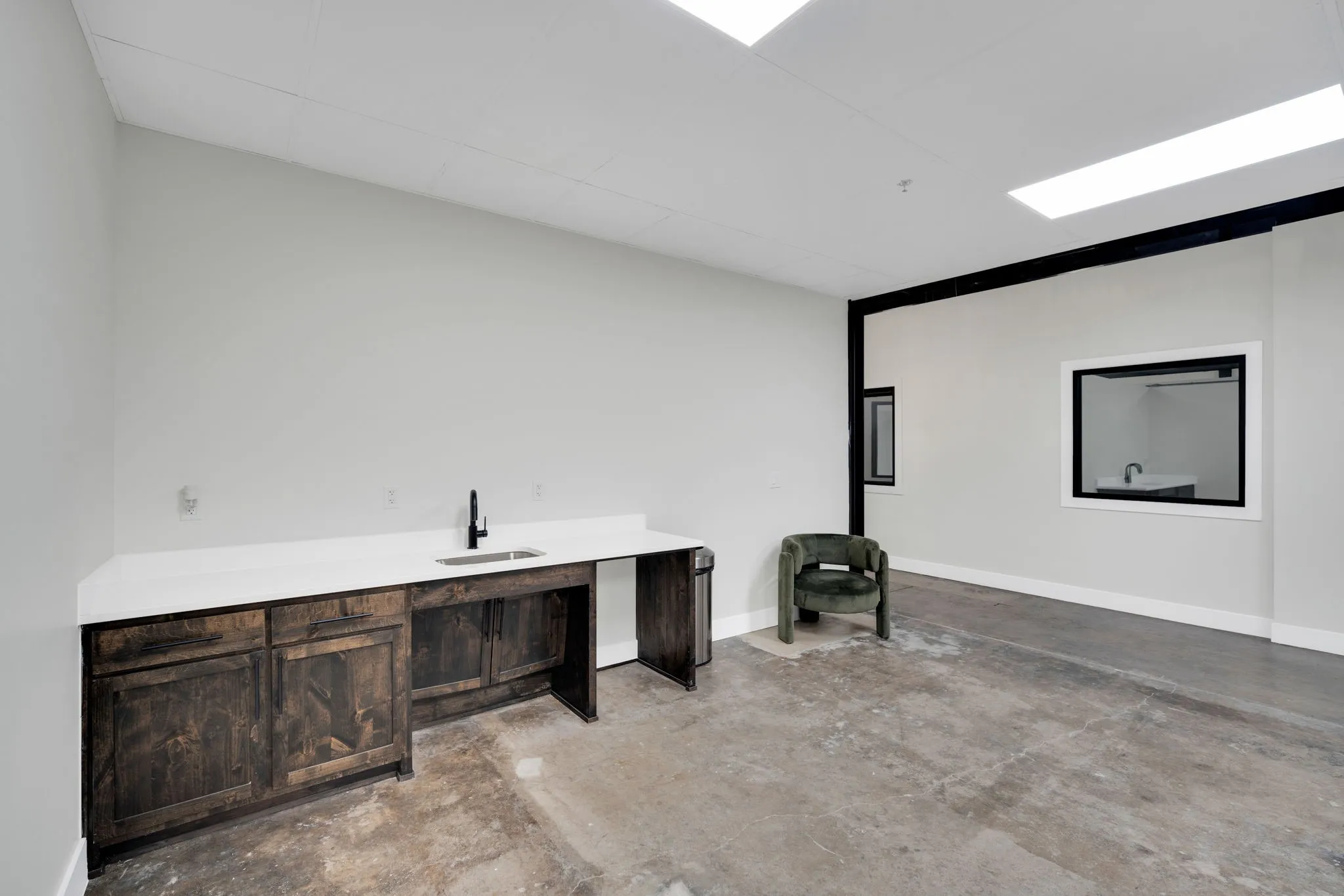 Spacious room featuring a built-in wood-finish cabinet with a white countertop and sink, light grey walls, and exposed concrete flooring