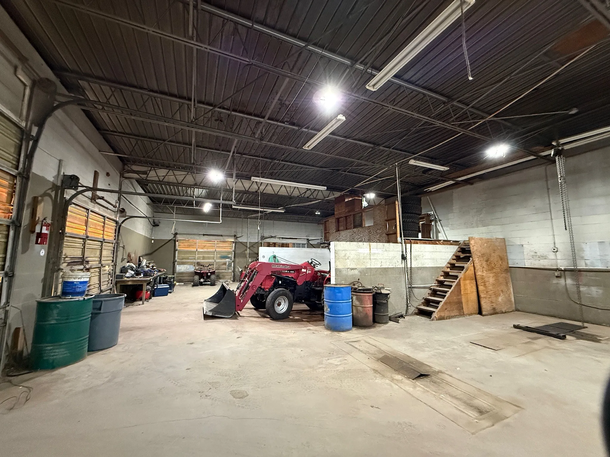 Spacious industrial interior featuring a high ceiling with exposed metal trusses, concrete flooring, multiple overhead fluorescent light fixtures, and a wooden mezzanine structure with a staircase