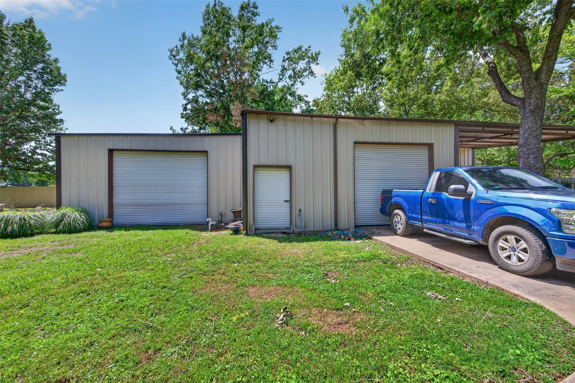 Metal-sided outbuilding featuring two garage doors, a pedestrian entrance, and an attached carport with a concrete driveway
