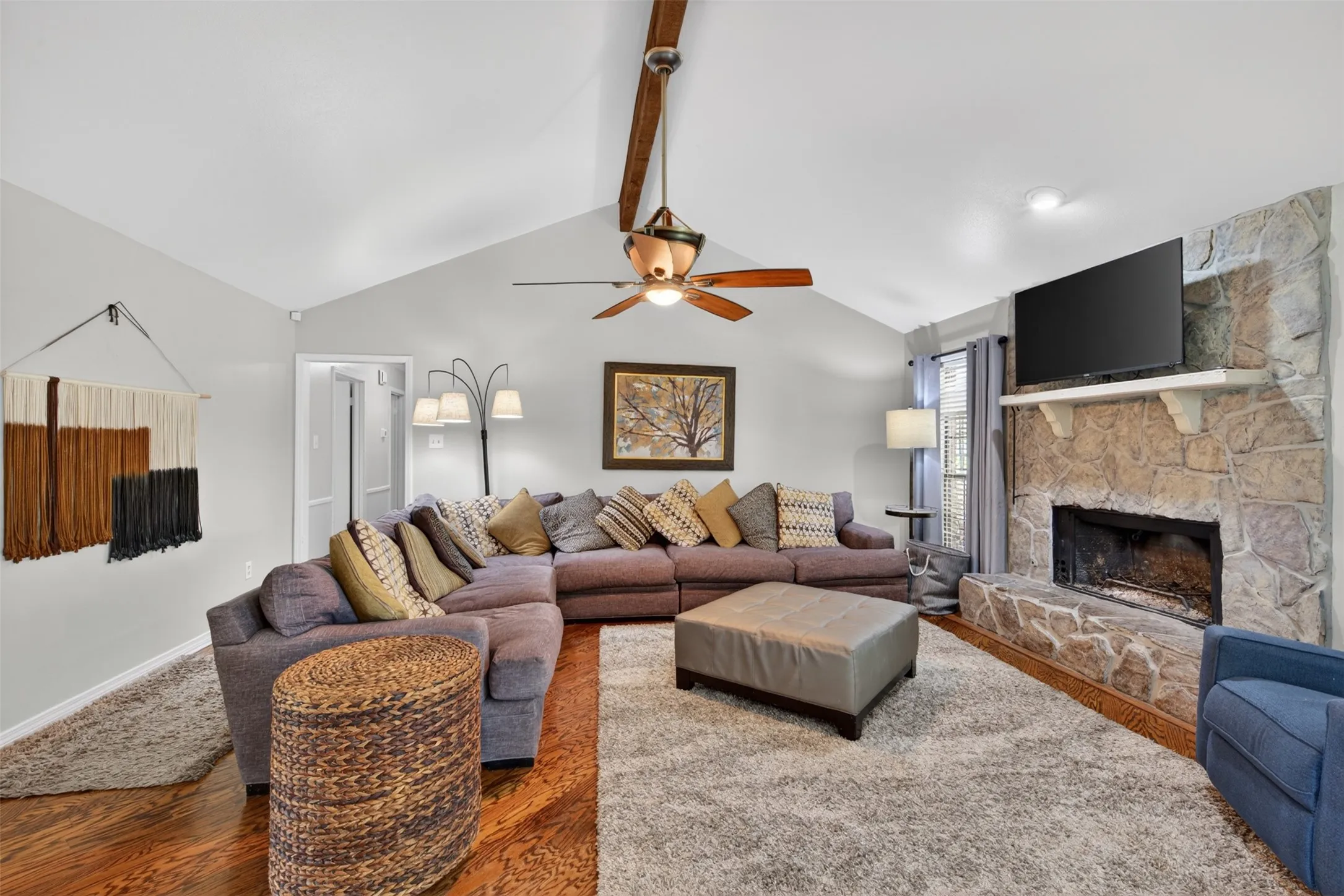 Spacious living area featuring a stone fireplace with a mantle, vaulted ceiling with a wood beam, and a ceiling fan