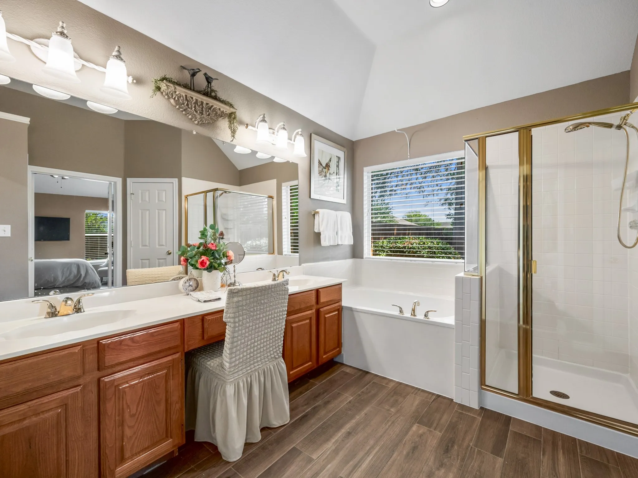 Bathroom featuring a double vanity with wood cabinetry, a built-in soaking tub, a separate glass-enclosed shower with gold-tone fixtures, and wood-finish flooring