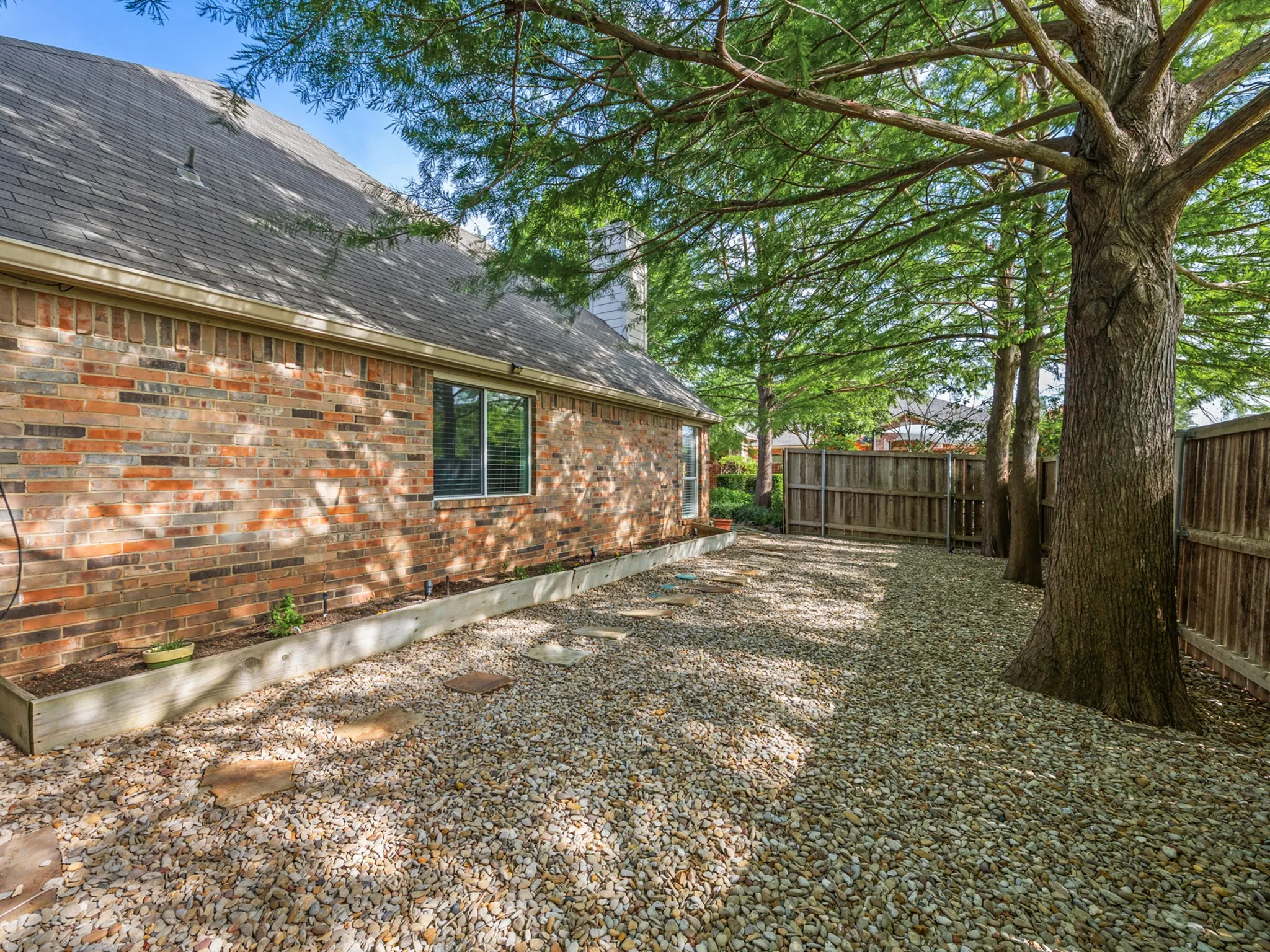 Gravel side yard with mature trees, brick exterior, wood fence, and raised planter bed