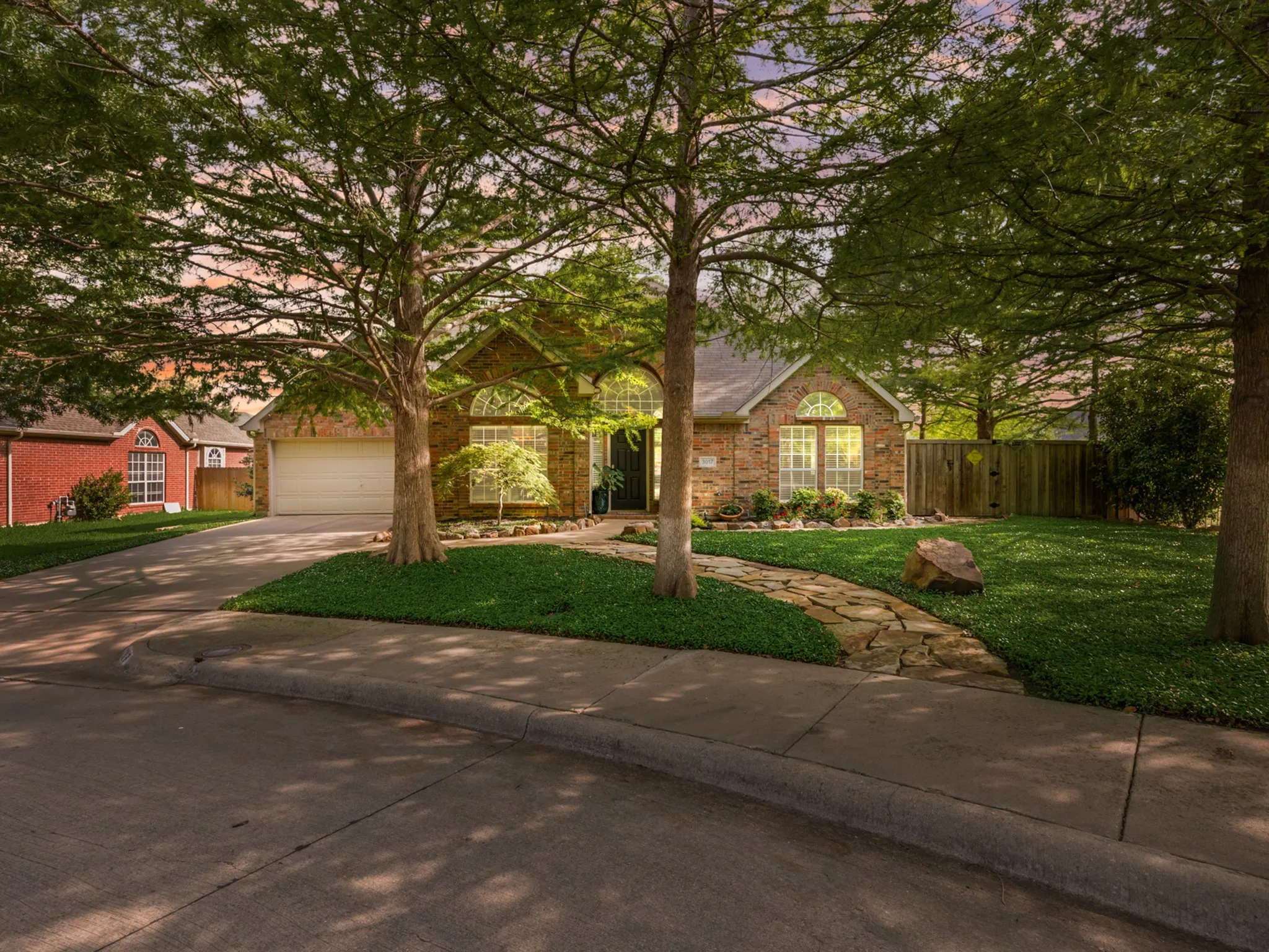 Brick-clad exterior featuring arched window details, a flagstone pathway, an attached two-car garage, mature trees, and a manicured lawn