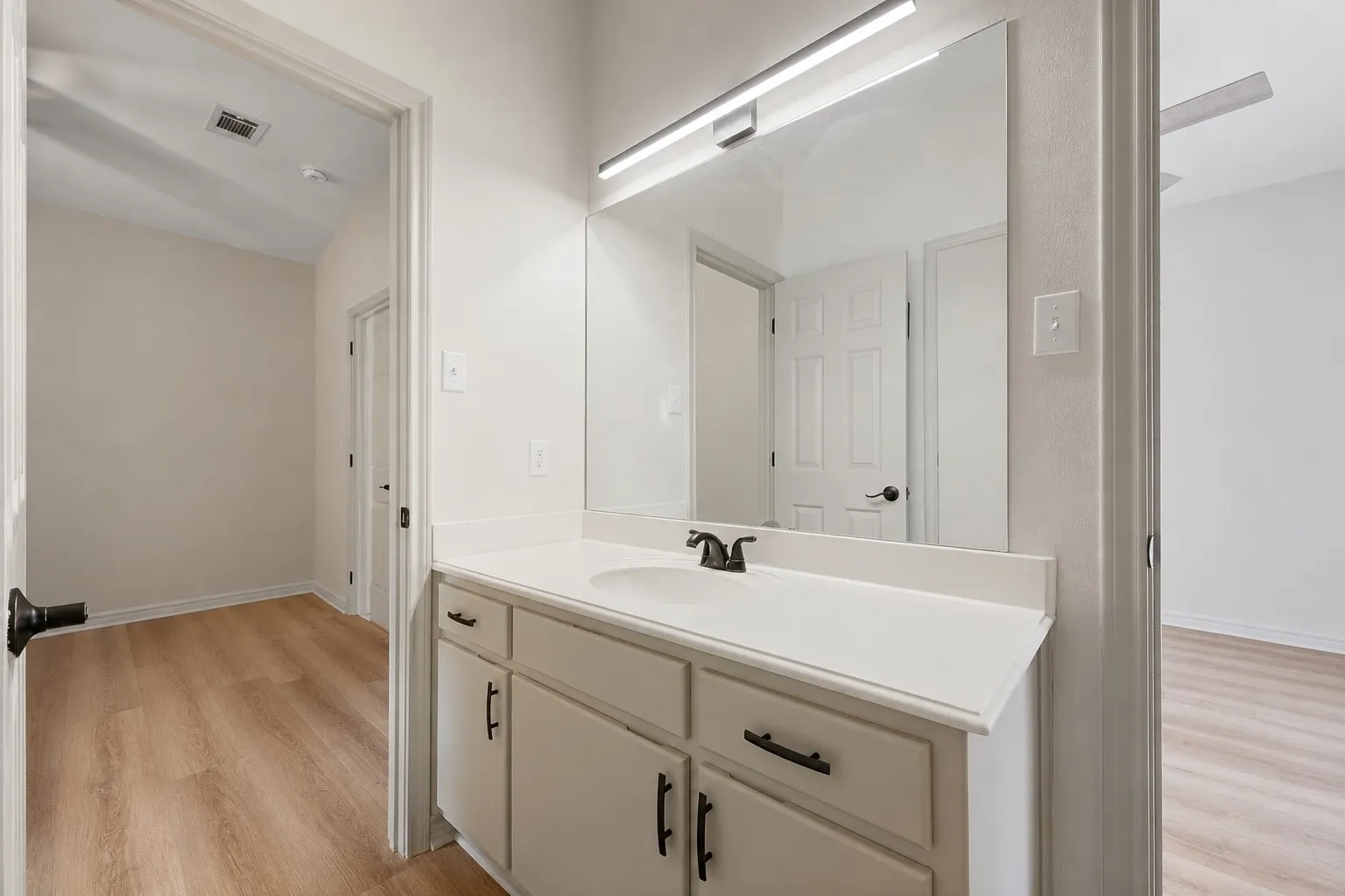Bathroom vanity featuring a single sink set into a light-toned countertop, white cabinetry with dark hardware, and a large mirror with integrated lighting