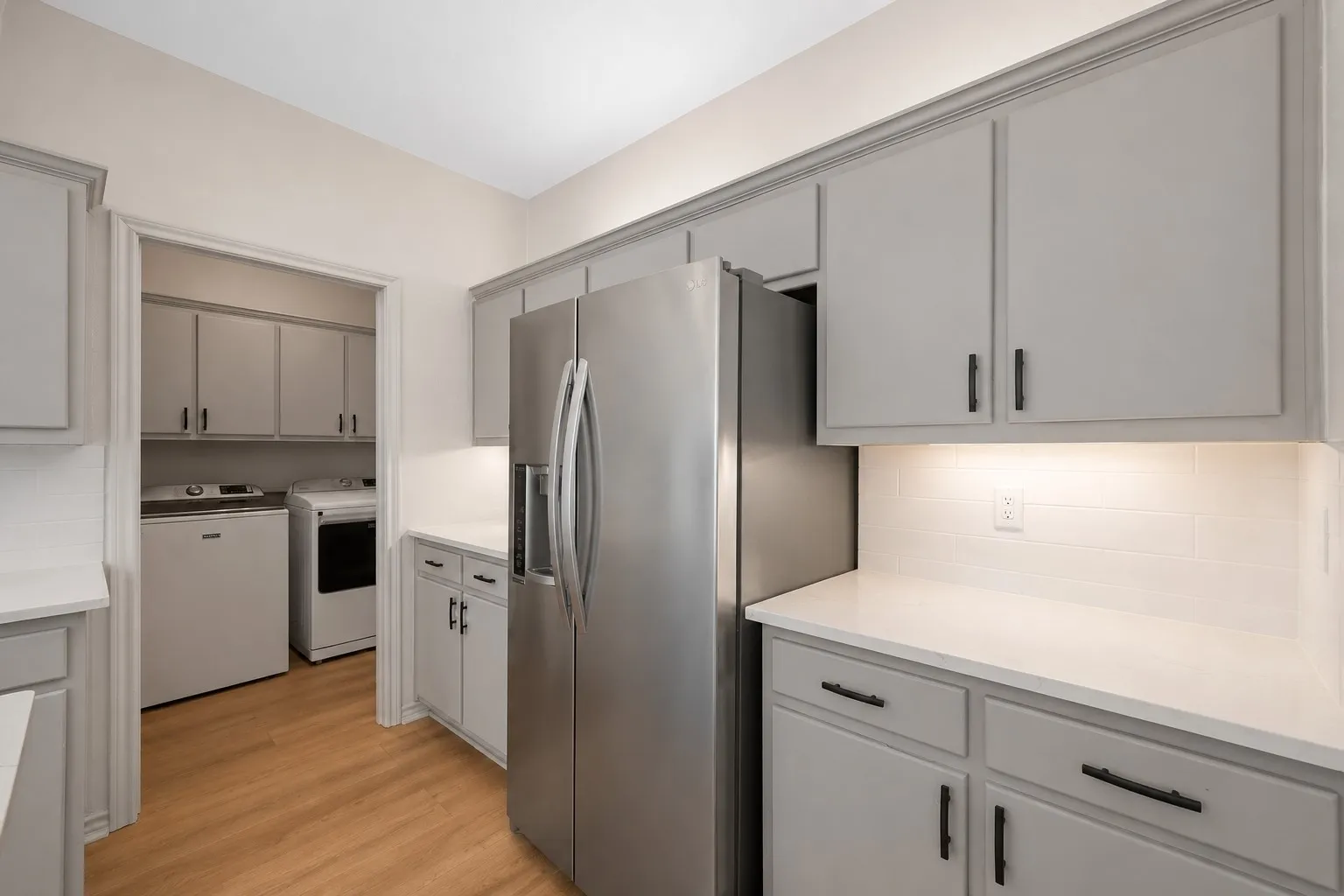 Kitchen area featuring light gray cabinetry, stainless steel refrigerator, white countertops, subway tile backsplash, and under-cabinet lighting