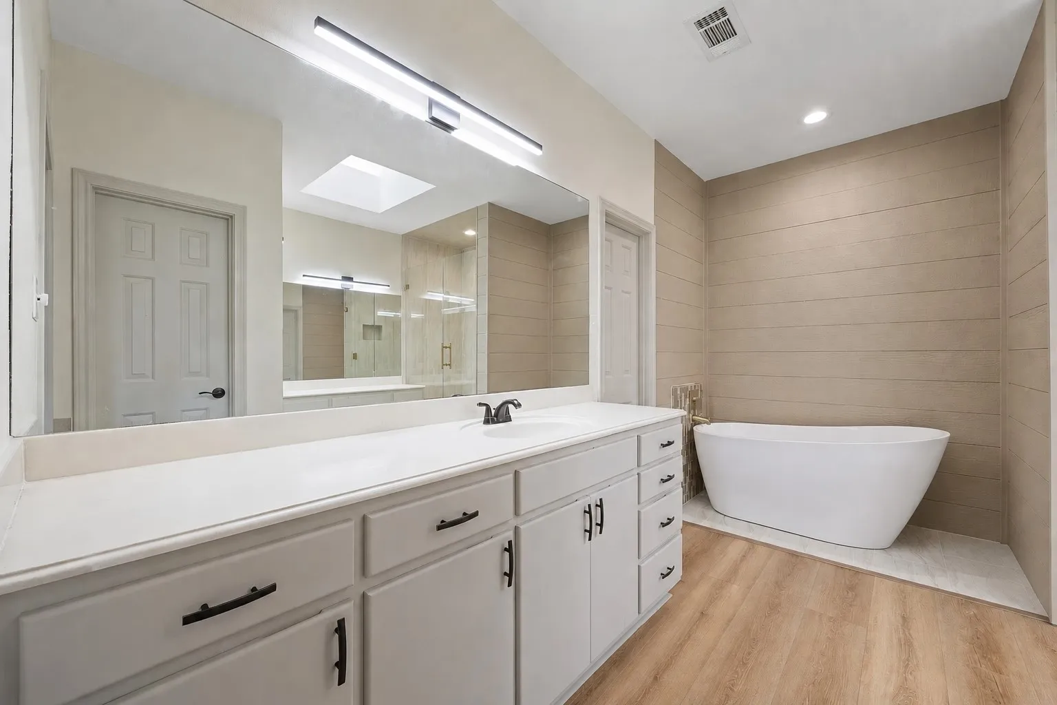 Primary Bathroom featuring a dual-sink vanity with white cabinetry, a freestanding soaking tub, wood-finish flooring, a large wall mirror, and a skylight