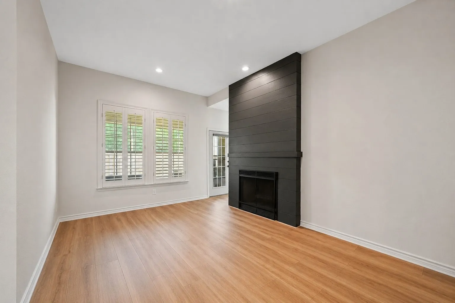 Living area featuring wood-finish flooring, a contemporary dark shiplap fireplace surround, recessed lighting, and white plantation shutters on a triple window unit