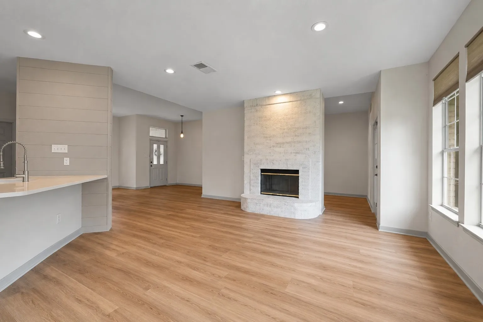 Open-concept living space with wood-finish flooring, a white brick fireplace, recessed lighting, and large windows with integrated blinds