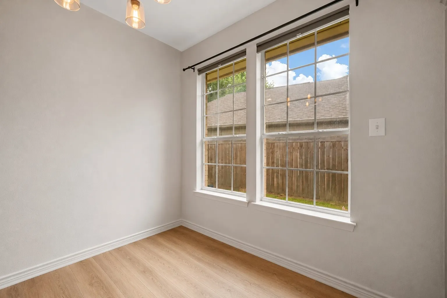 Room featuring two large windows with white frames, wood-finish flooring, light neutral wall paint, white baseboards, and a contemporary ceiling light fixture with amber glass shades