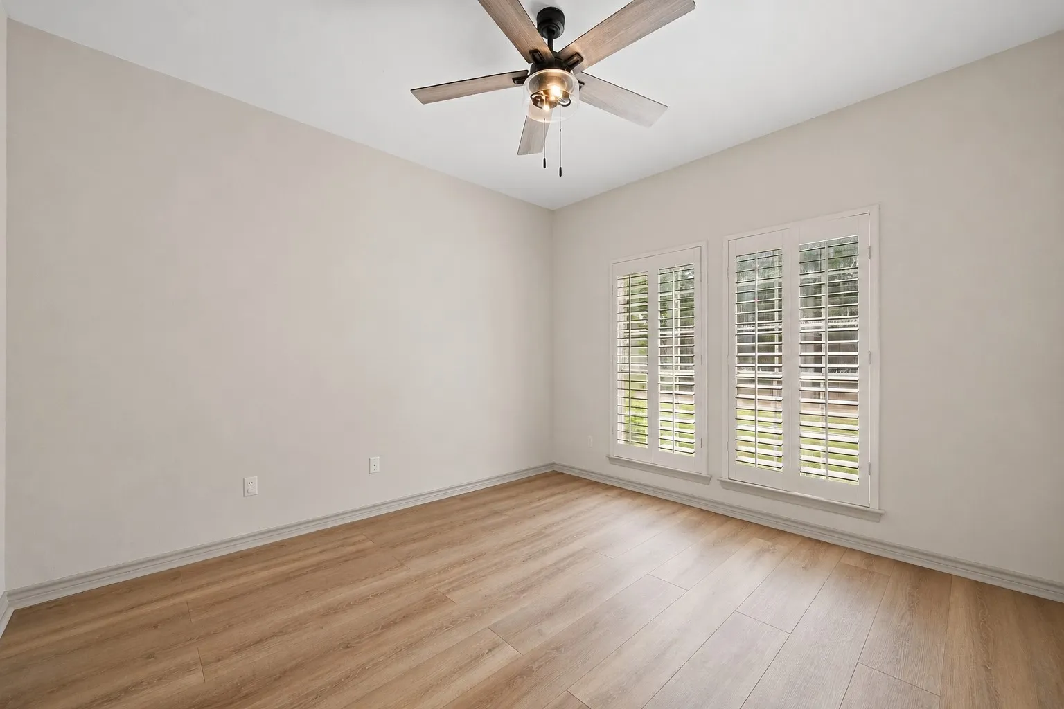 Room featuring light wood-finish flooring, two large windows with white louvered shutters, and a ceiling fan with integrated lighting