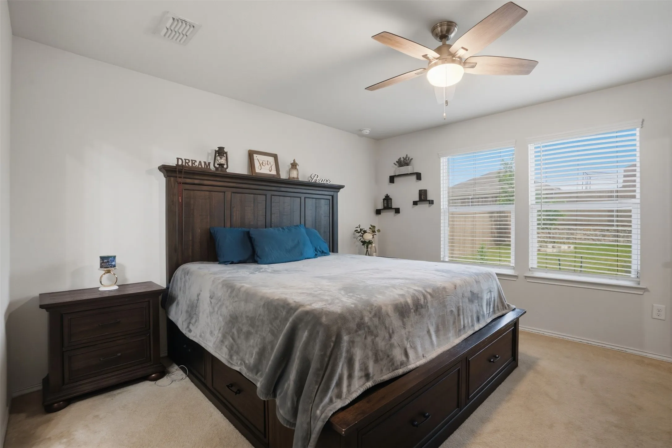 Carpeted room featuring a ceiling fan with integrated lighting, two windows with horizontal blinds, and built-in floating shelves