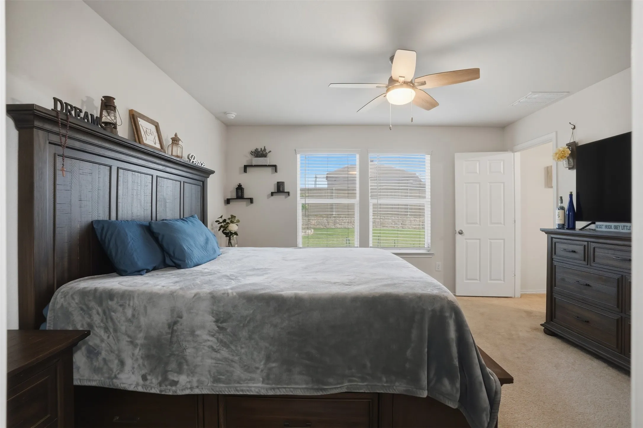 Carpeted bedroom featuring a ceiling fan, two windows with blinds, a paneled door, and built-in wall shelving