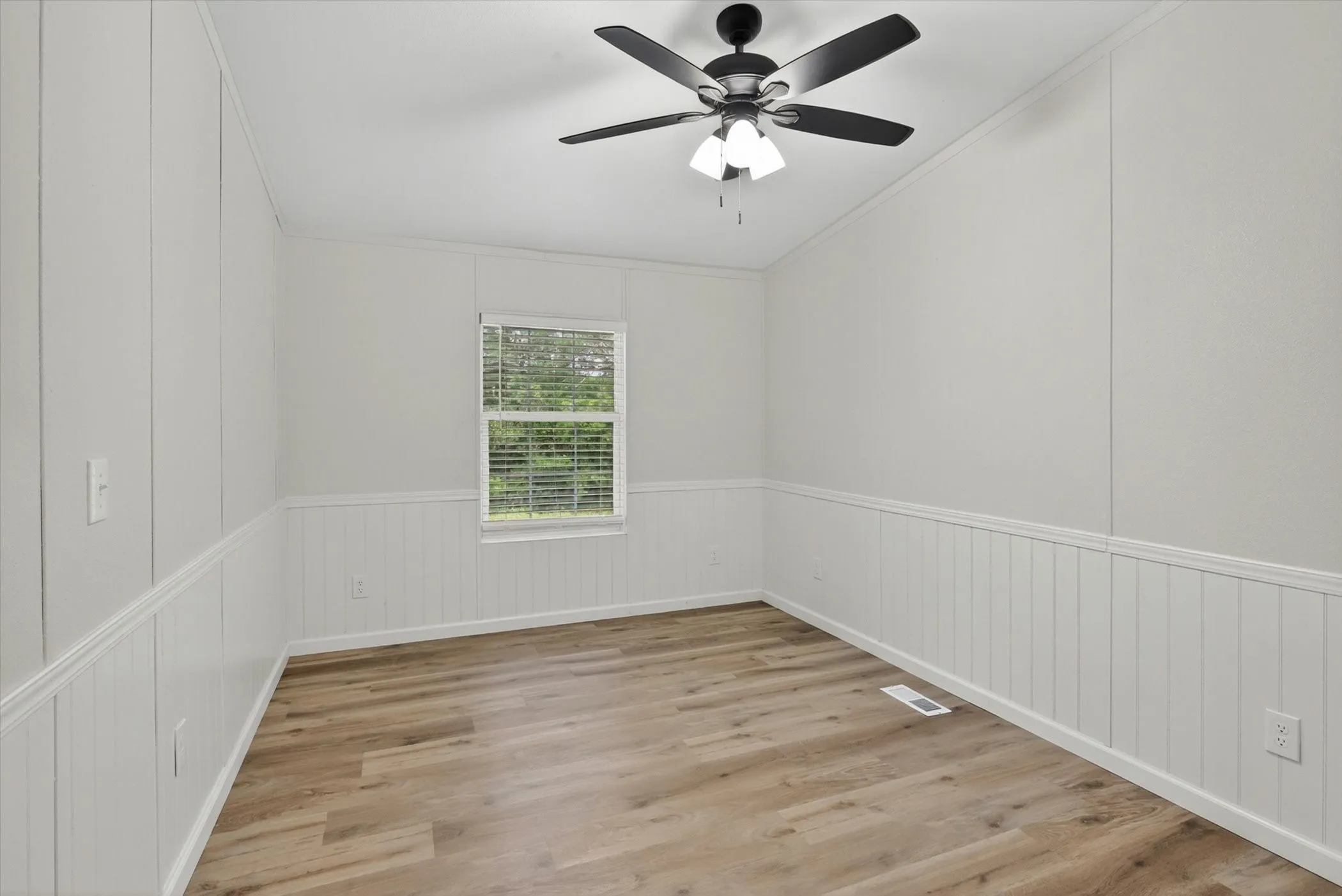 Spacious room featuring wood-finish flooring, white wainscoting, and a ceiling fan with integrated lighting