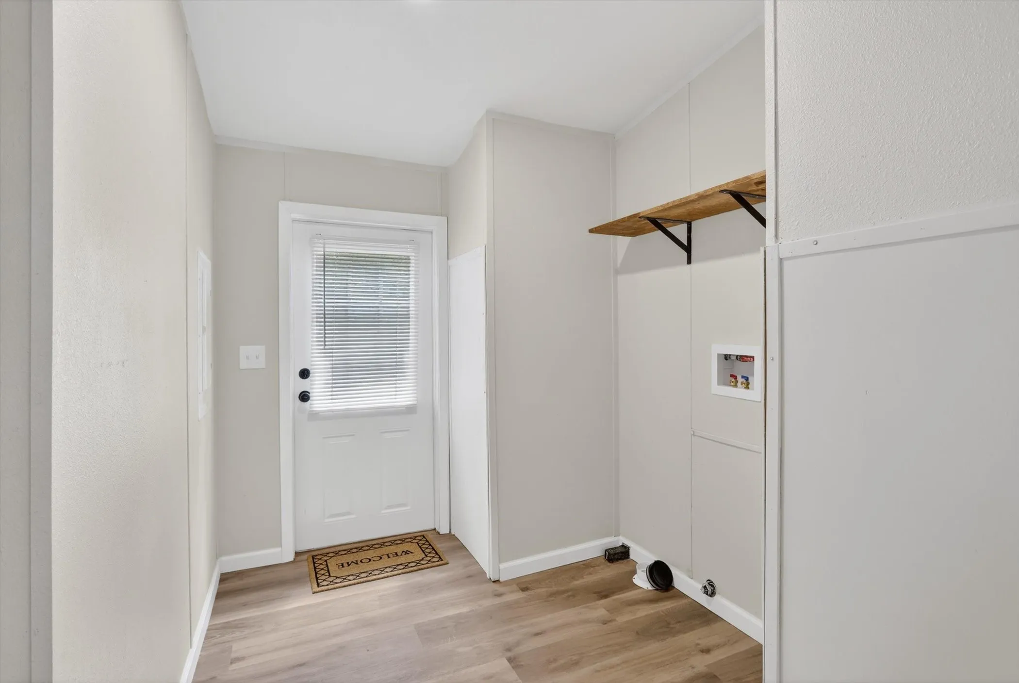 Utility room featuring wood-finish flooring, a white paneled door with integrated blinds, and a built-in wood shelf with black brackets