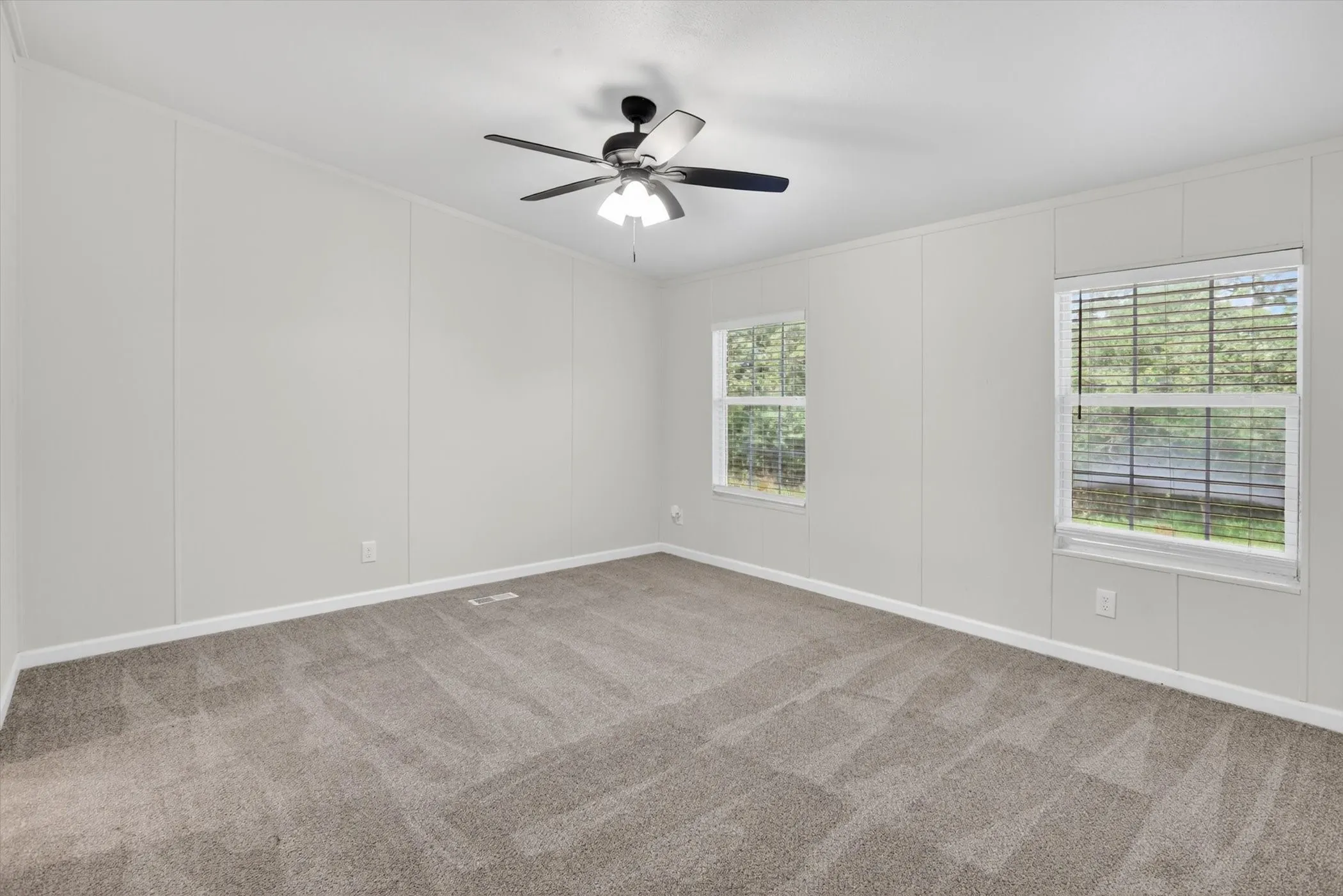 Spacious room featuring neutral carpeting, two windows with blinds, a ceiling fan with integrated lighting, and light-toned wall paneling