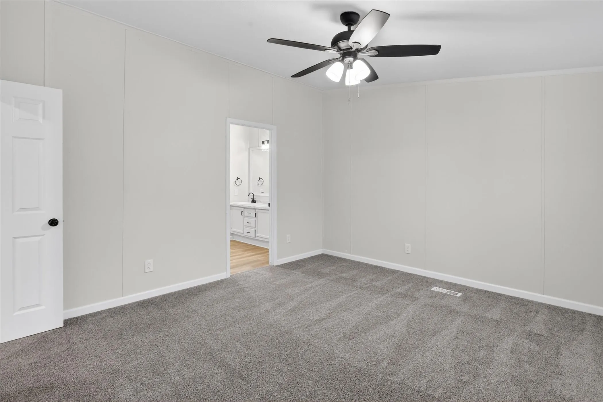 Spacious room featuring gray carpet flooring, light gray walls, a white paneled door with black hardware, and a ceiling fan with light fixture