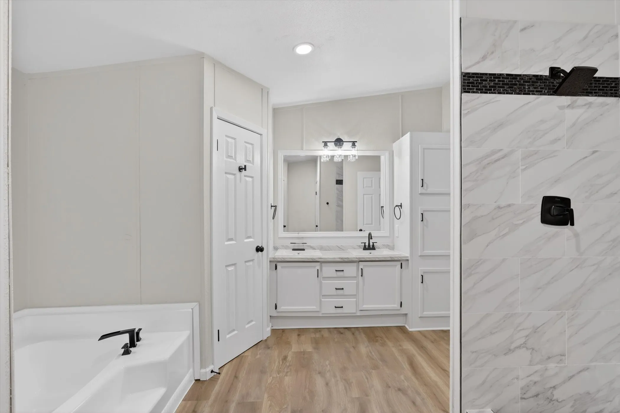 Bathroom featuring a dual vanity, white cabinetry, wood-finish flooring, a soaking tub with dark fixtures, and a shower with tile surround and dark fixtures