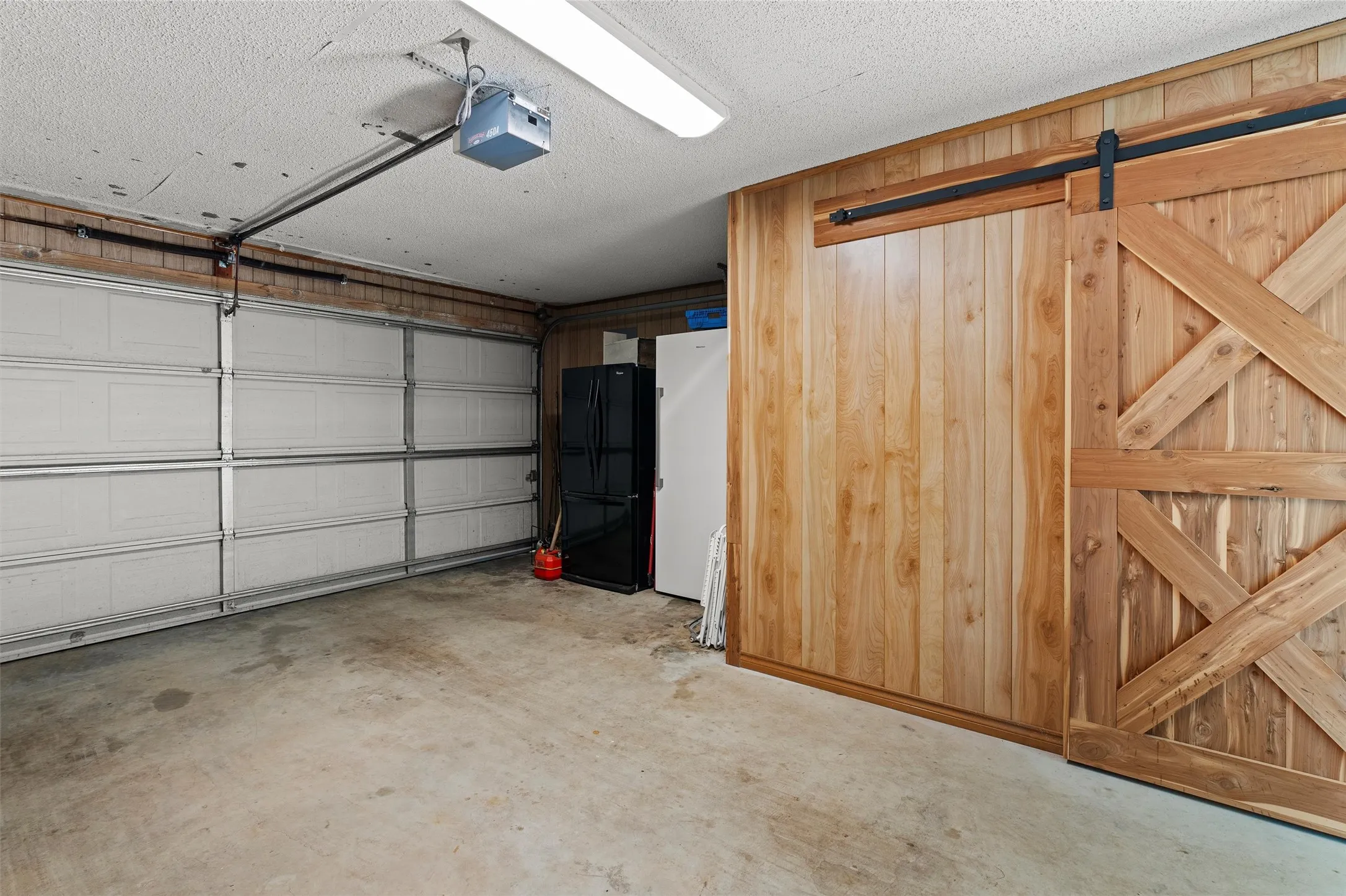 Spacious garage featuring a concrete floor, a white paneled garage door, a garage door opener, wood-paneled walls, and a wood barn door on a black metal track