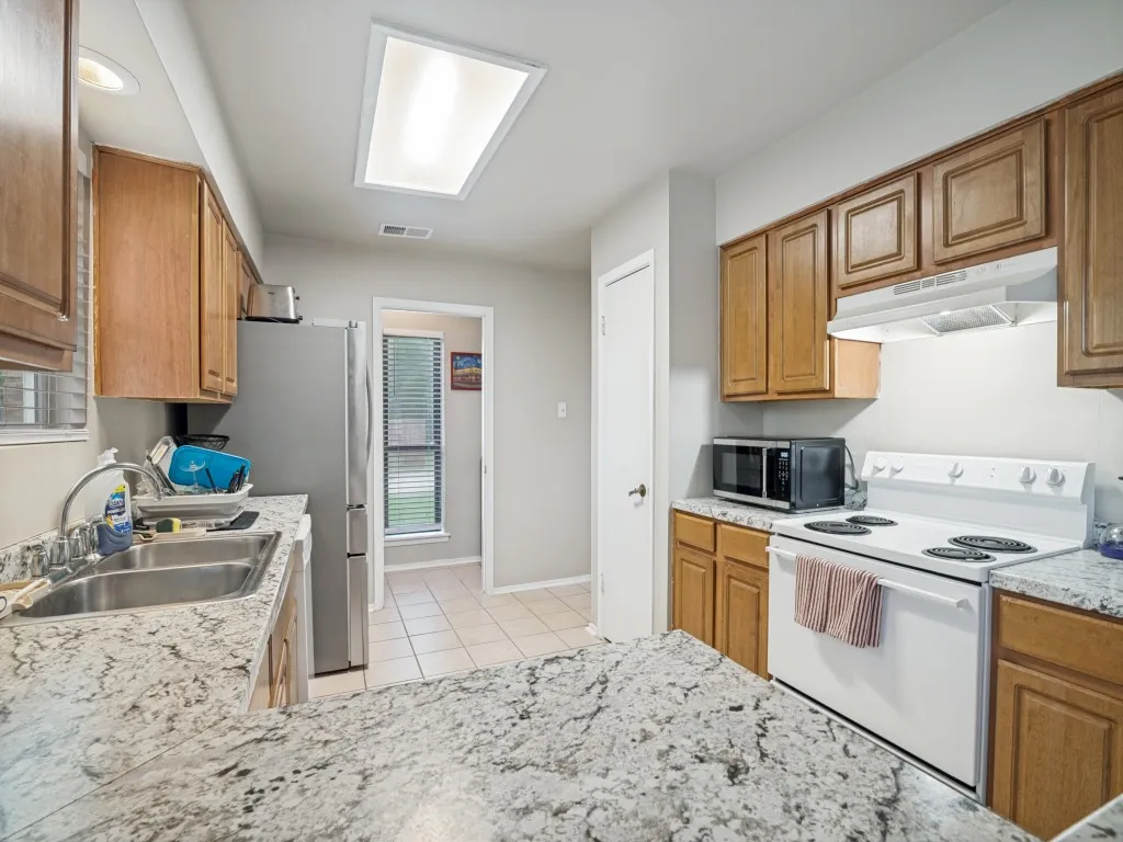 Spacious kitchen featuring light wood cabinetry, stone-finish countertops, stainless steel double basin sink, and a large recessed ceiling light fixture