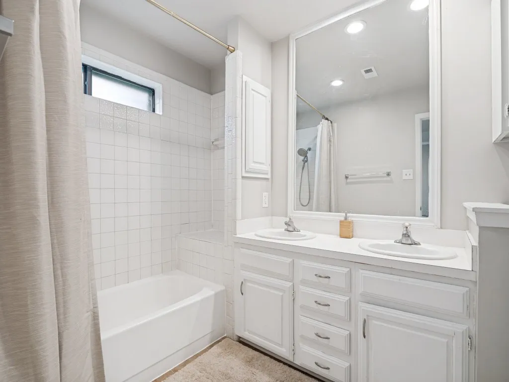 Bathroom featuring a dual-sink vanity with white cabinetry, a built-in bathtub with white tile surround, and a frosted window for natural light