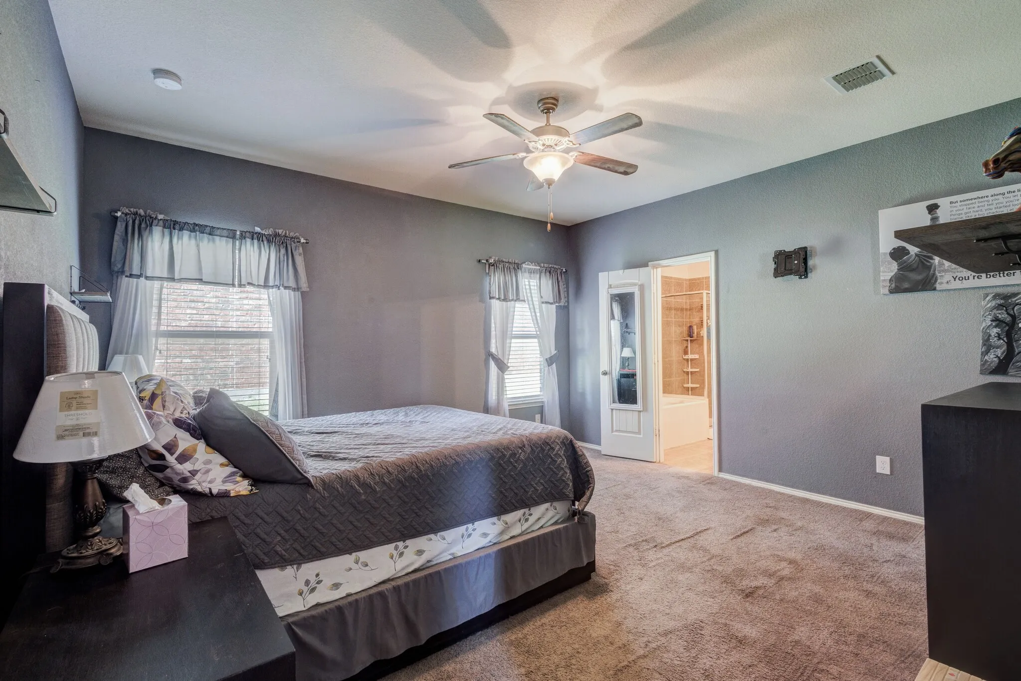 Carpeted bedroom with a ceiling fan, two windows with blinds, and an interior door revealing a bathroom with a tub/shower combination