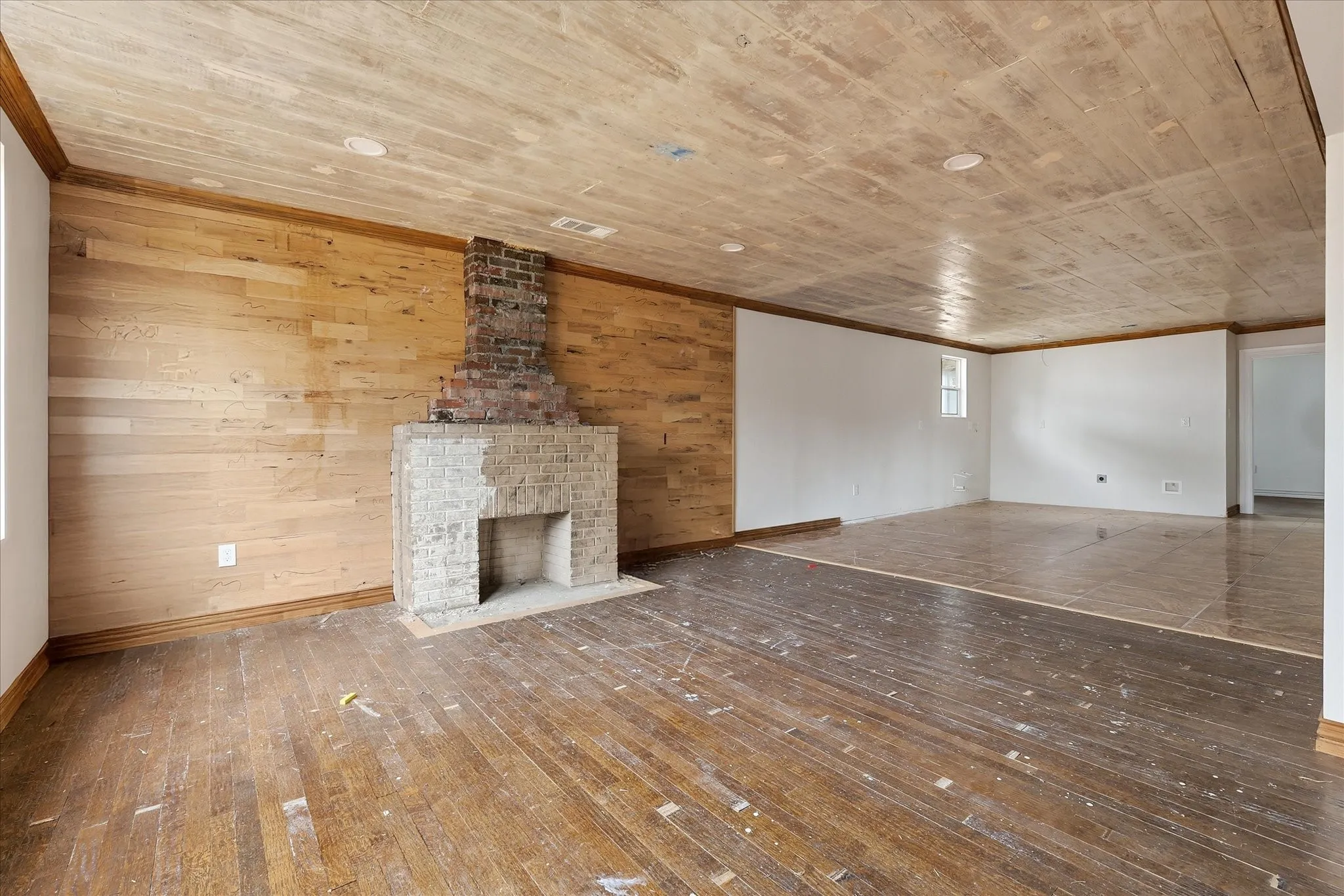 Spacious room featuring a brick fireplace with a light-colored brick hearth, wood-paneled accent wall, wood-finish flooring, recessed lighting, and a white-painted window
