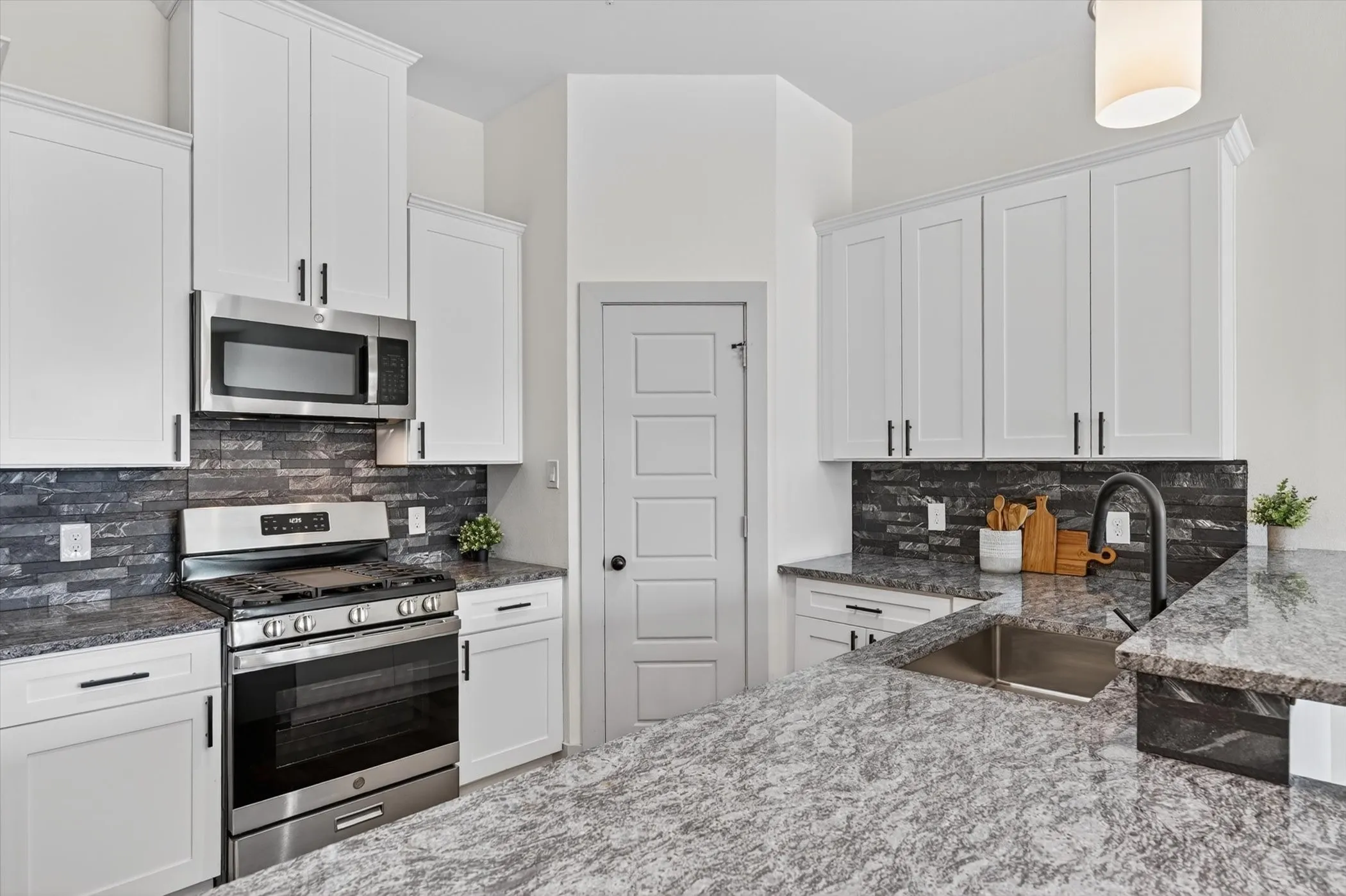 Modern kitchen featuring white shaker cabinetry, stainless steel appliances, a dark stacked stone backsplash, and light granite countertops with an undermount sink
