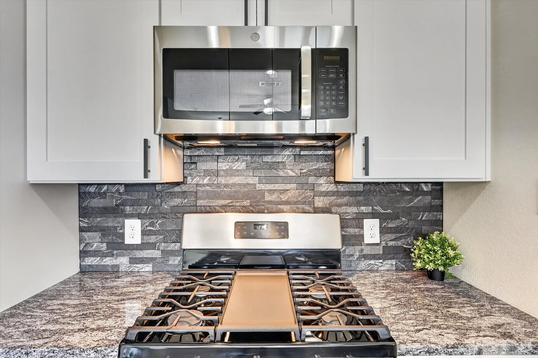 Kitchen featuring a gas range, a mounted stainless steel microwave, stone tile backsplash, light-toned cabinetry with dark pulls, and a speckled countertop