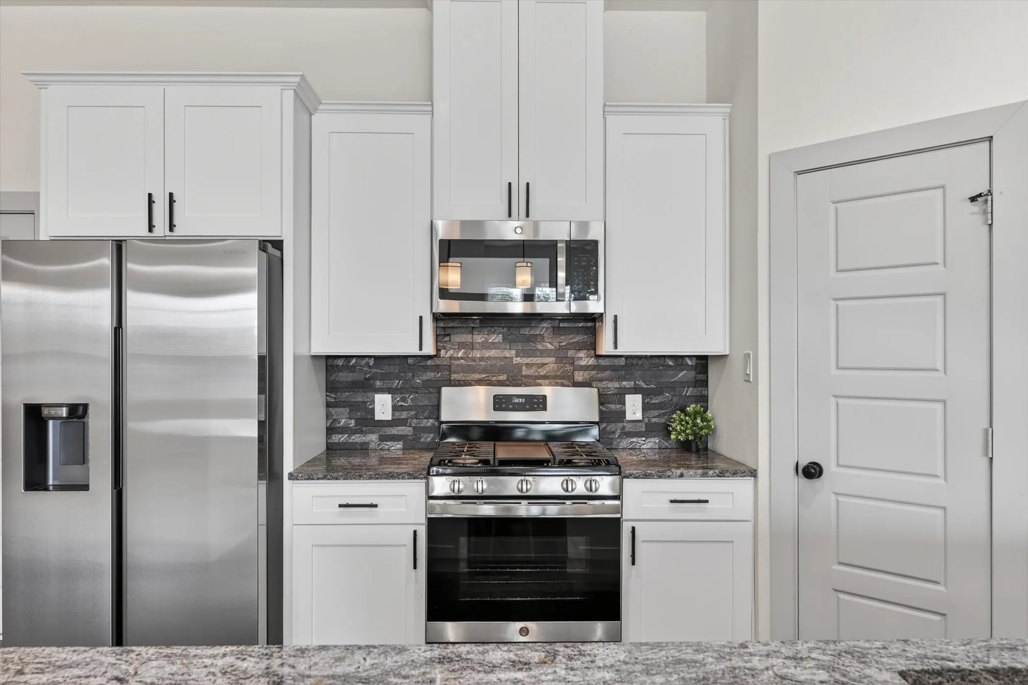 Modern kitchen featuring stainless steel appliances, white shaker cabinetry with black hardware, a dark stacked stone backsplash, and granite countertops