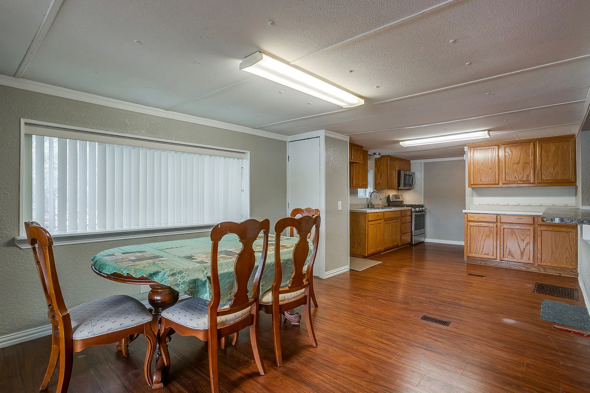 Open-concept living space featuring wood-finish flooring, a large window with vertical blinds, and a kitchen with wood cabinetry and light countertops