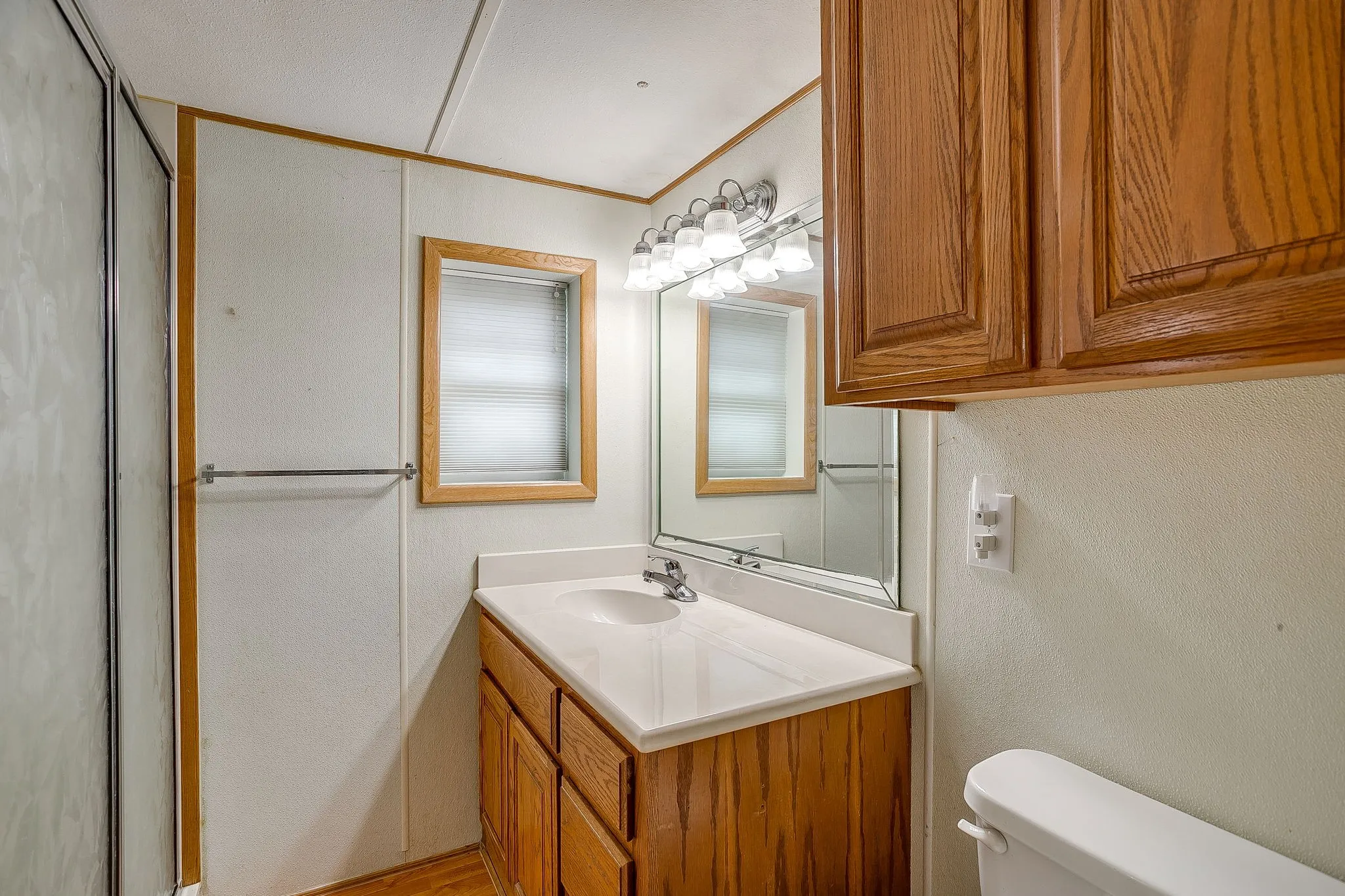 Bathroom featuring a wooden vanity with integrated sink, a large wall-mounted mirror, overhead lighting with frosted glass shades, a window with horizontal blinds, and a shower enclosure with frosted glass