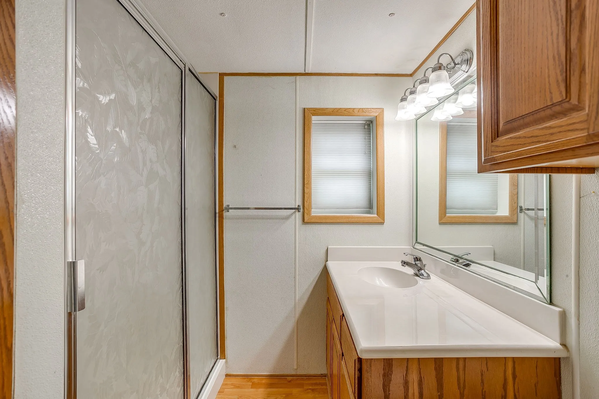 Bathroom featuring a shower stall with patterned glass, a single vanity with an integrated sink, an overhead light fixture with five bulbs, a framed mirror, and wood-finish flooring