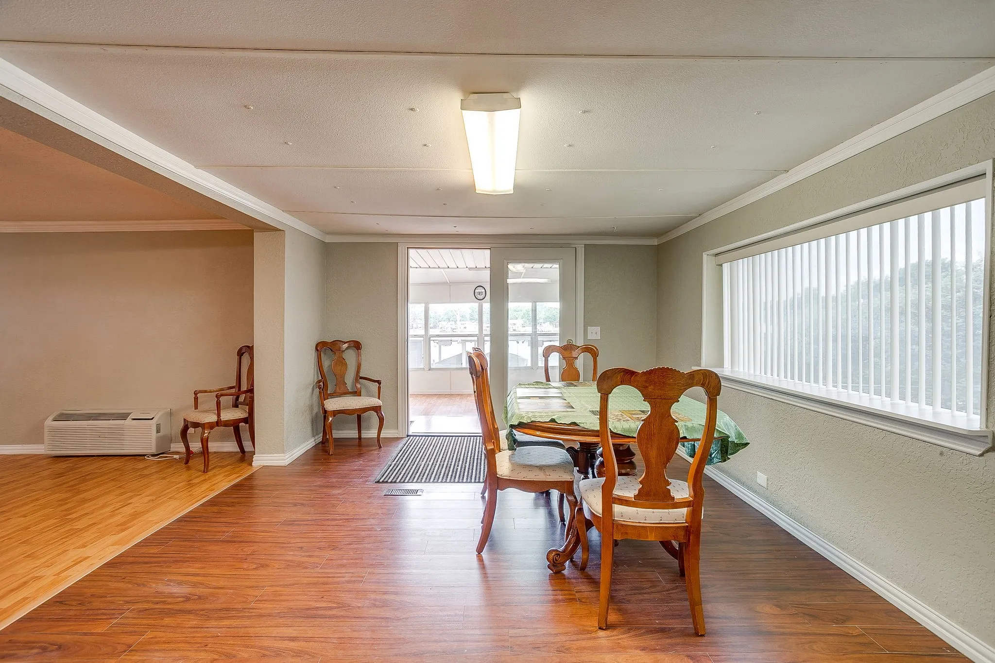 Spacious room featuring wood-finish flooring, a broad window with vertical blinds, and direct access to an enclosed sunroom