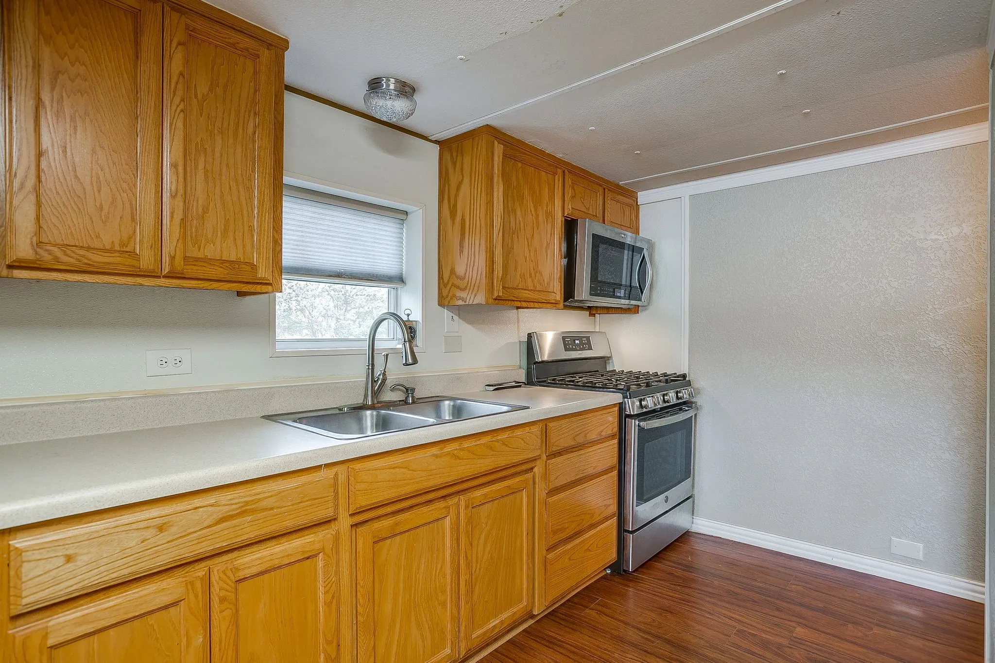 Kitchen featuring wood cabinetry, light-toned countertops, a double basin stainless steel sink with a gooseneck faucet, stainless steel microwave, and a stainless steel gas range